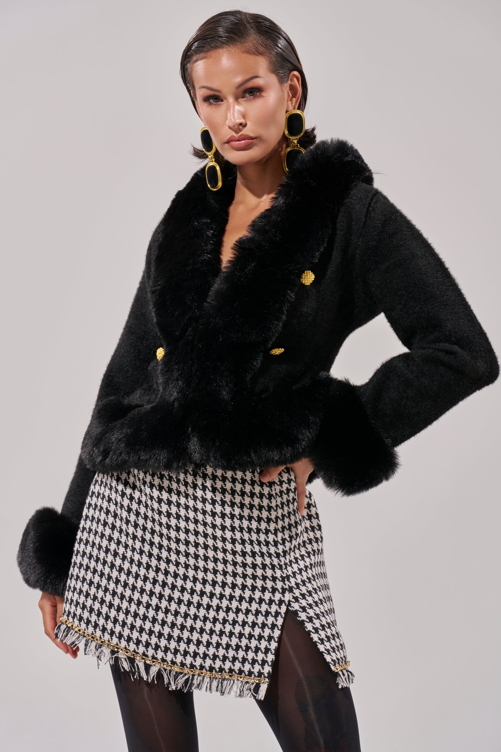 A woman poses in the SOFT TOUCH CARDIGAN IN BLACK, styled with a black-and-white houndstooth mini skirt, black tights, and gold hoop earrings—an elegant choice for any winter wardrobe.