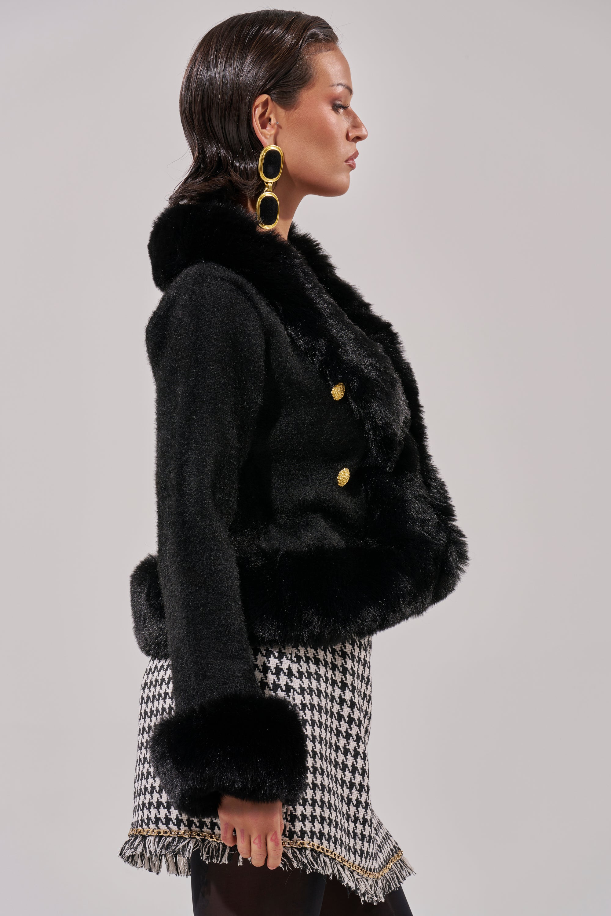 A woman in profile wears the SOFT TOUCH CARDIGAN IN BLACK with gold buttons, styled with large gold statement earrings and a black-and-white houndstooth fringe skirt, standing against a plain light background.
