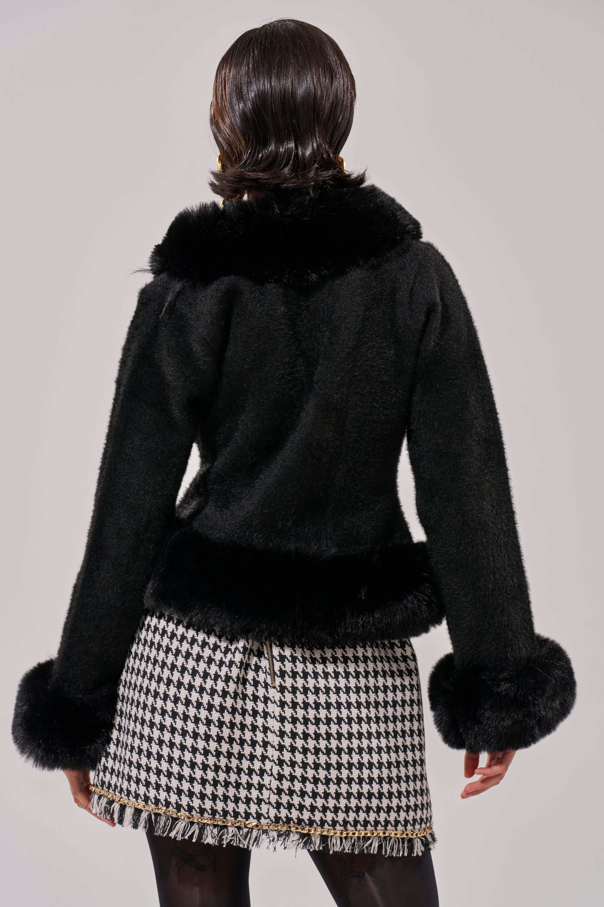 A person is seen from behind wearing the SOFT TOUCH CARDIGAN IN BLACK, featuring faux fur trim with large cuffs and collar, paired with a black-and-white houndstooth skirt with a fringe hem against a plain neutral background.