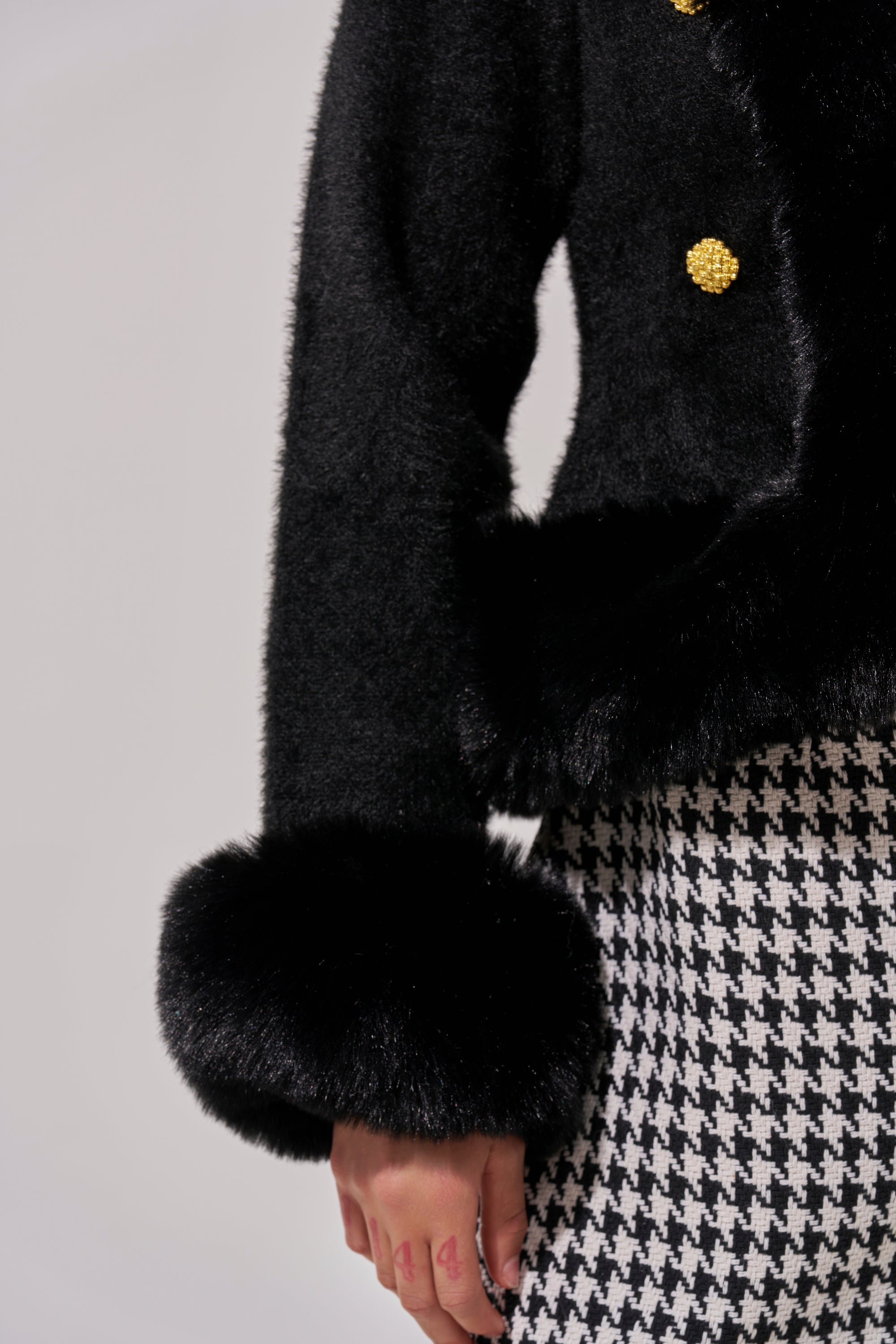 A person wearing the SOFT TOUCH CARDIGAN IN BLACK with gold buttons and thick faux fur cuffs, paired with black and white houndstooth pants; only the lower part of the cardigan and one hand are visible.