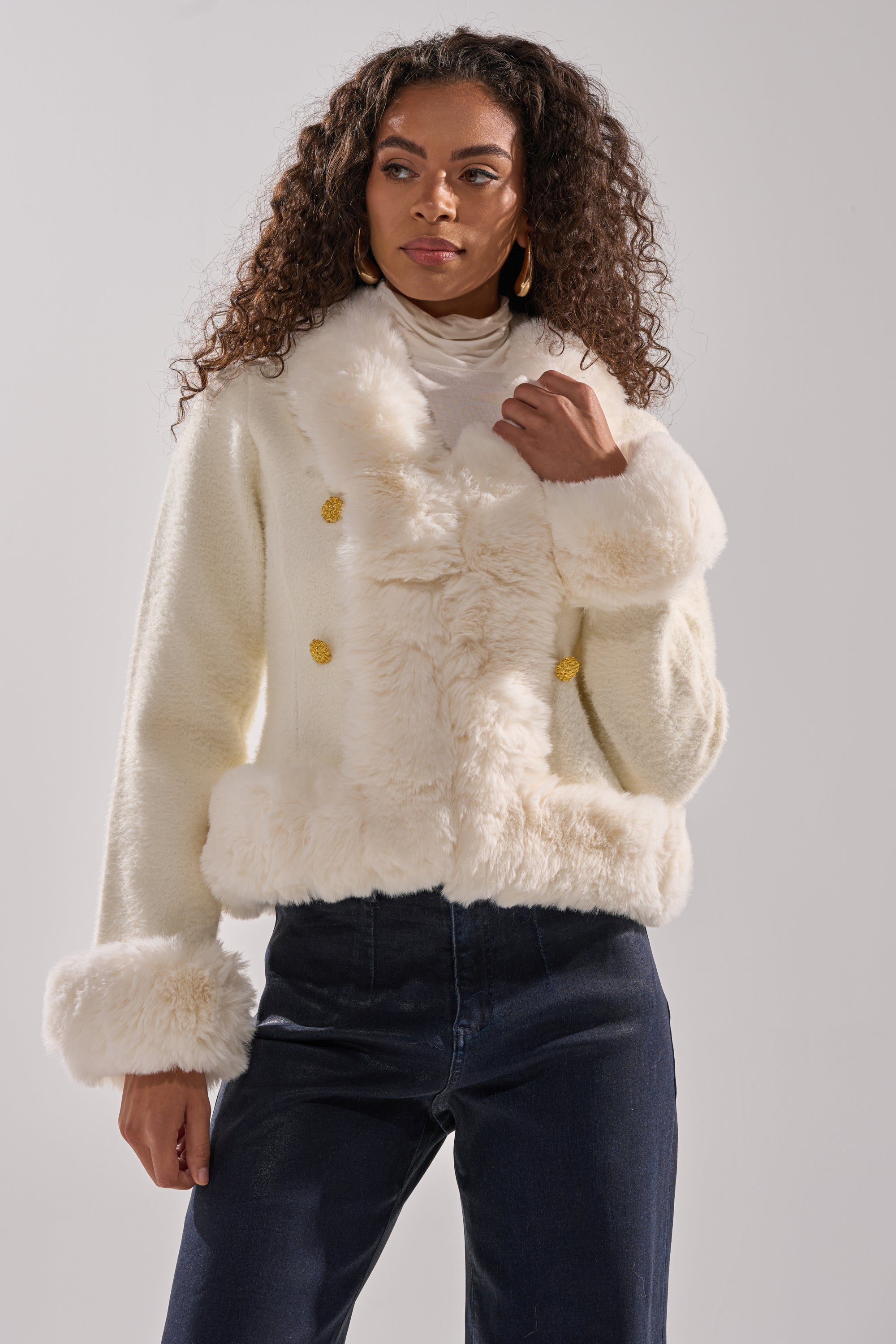 A woman with long curly hair wears the SOFT TOUCH CARDIGAN, layered under a white faux fur jacket with gold buttons, paired with a white top and dark blue high-waisted pants against a plain light background. 0