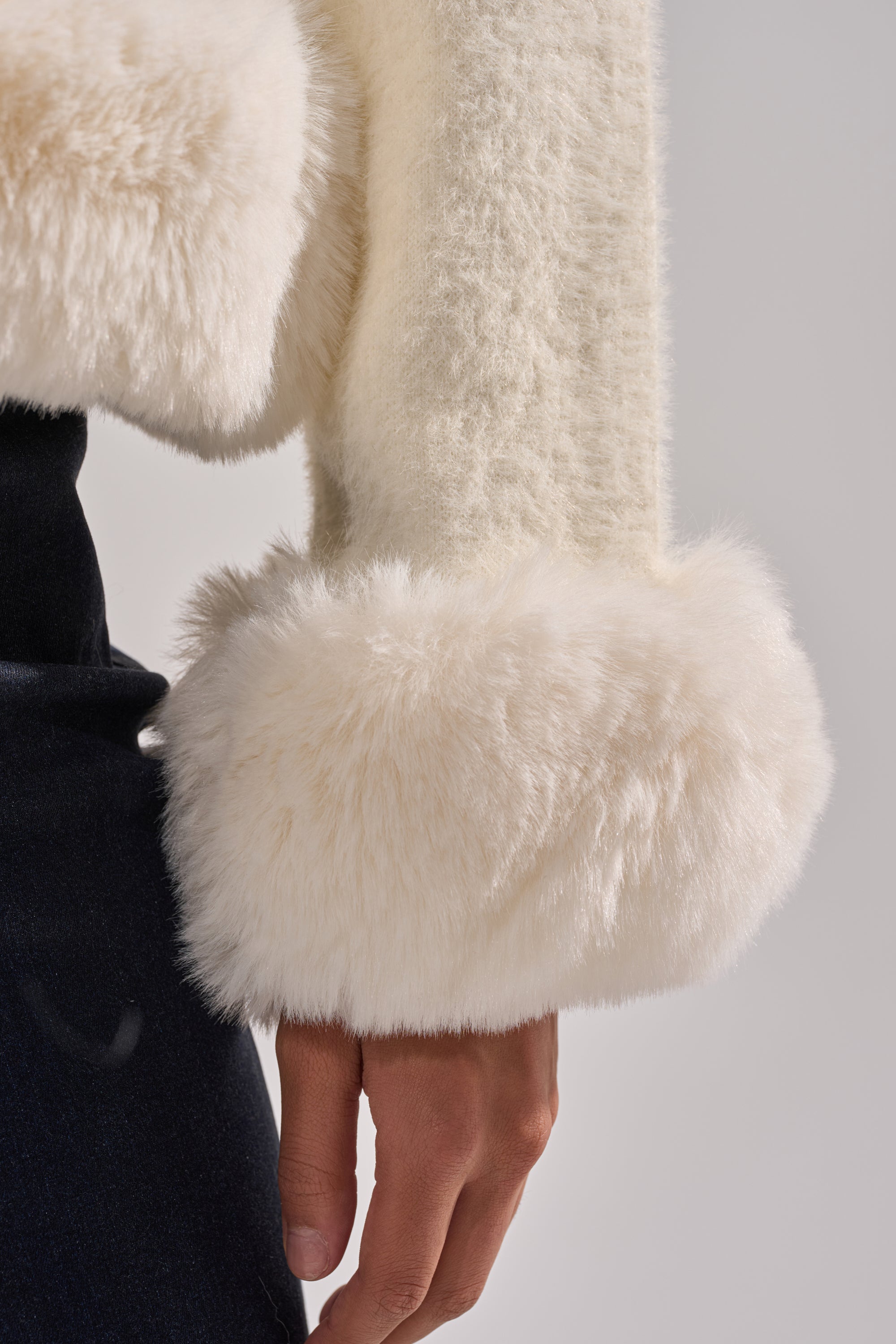 A close-up of a person's arm in the SOFT TOUCH CARDIGAN, featuring a cream knit with thick, fluffy faux fur trim cuffs, paired with dark clothing for a soft and cozy look. 9