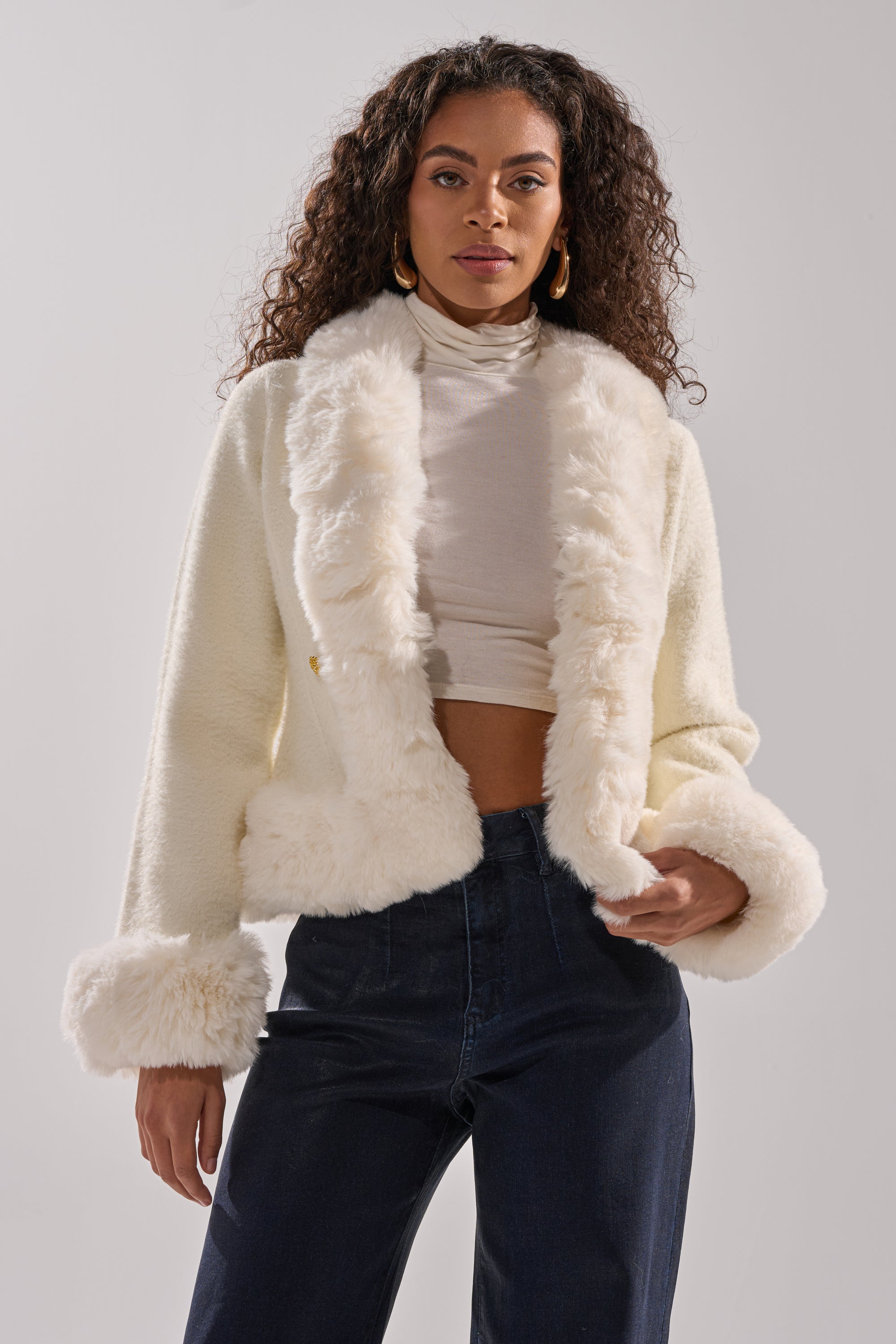 A woman with long curly hair wears the SOFT TOUCH CARDIGAN over a white turtleneck crop top and high-waisted dark jeans, standing against a plain light gray background. 1