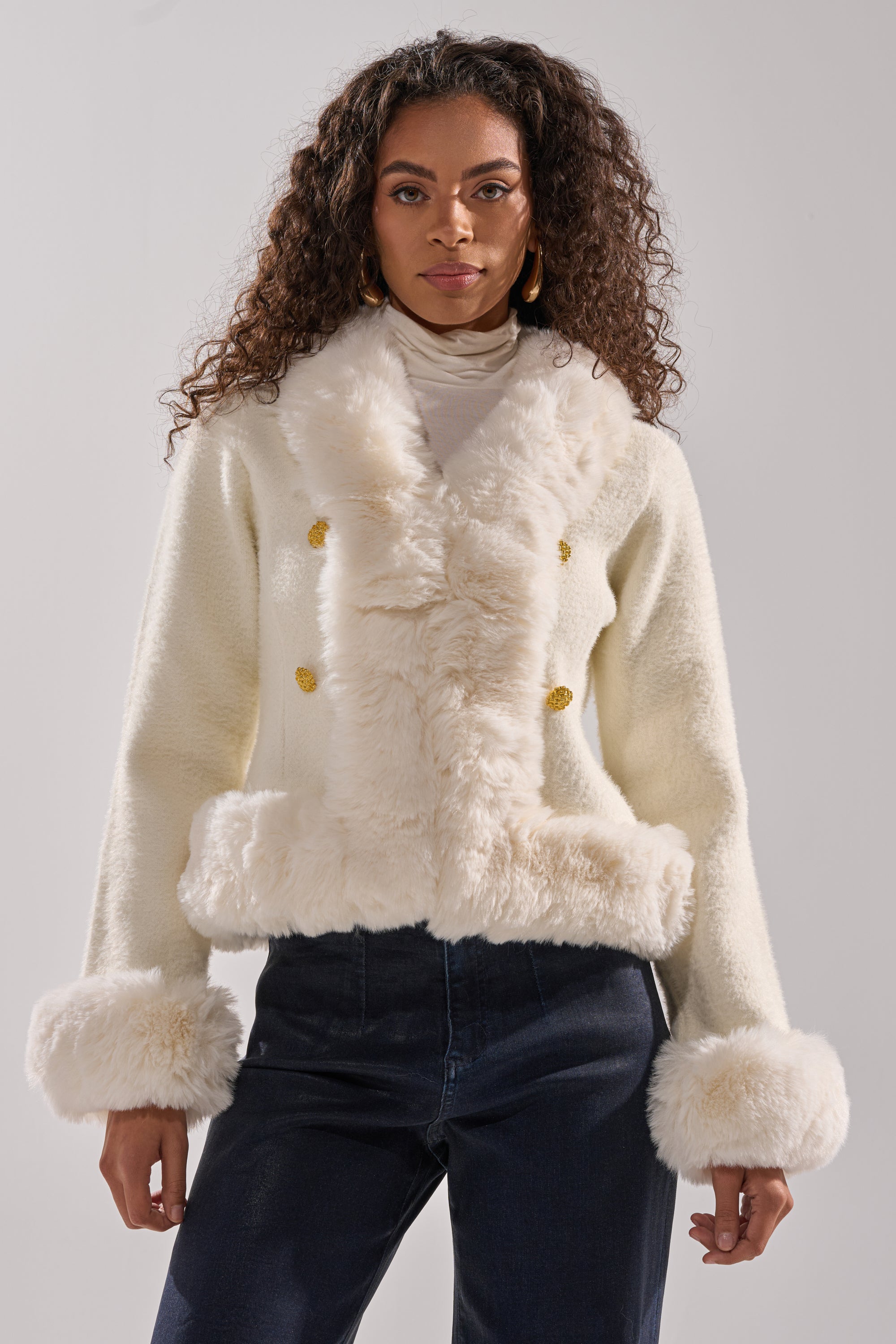 A woman with long curly hair wears the SOFT TOUCH CARDIGAN, featuring white faux fur trim, gold buttons, and furry cuffs and hem over a light turtleneck and dark pants. She stands against a plain, light background. 2