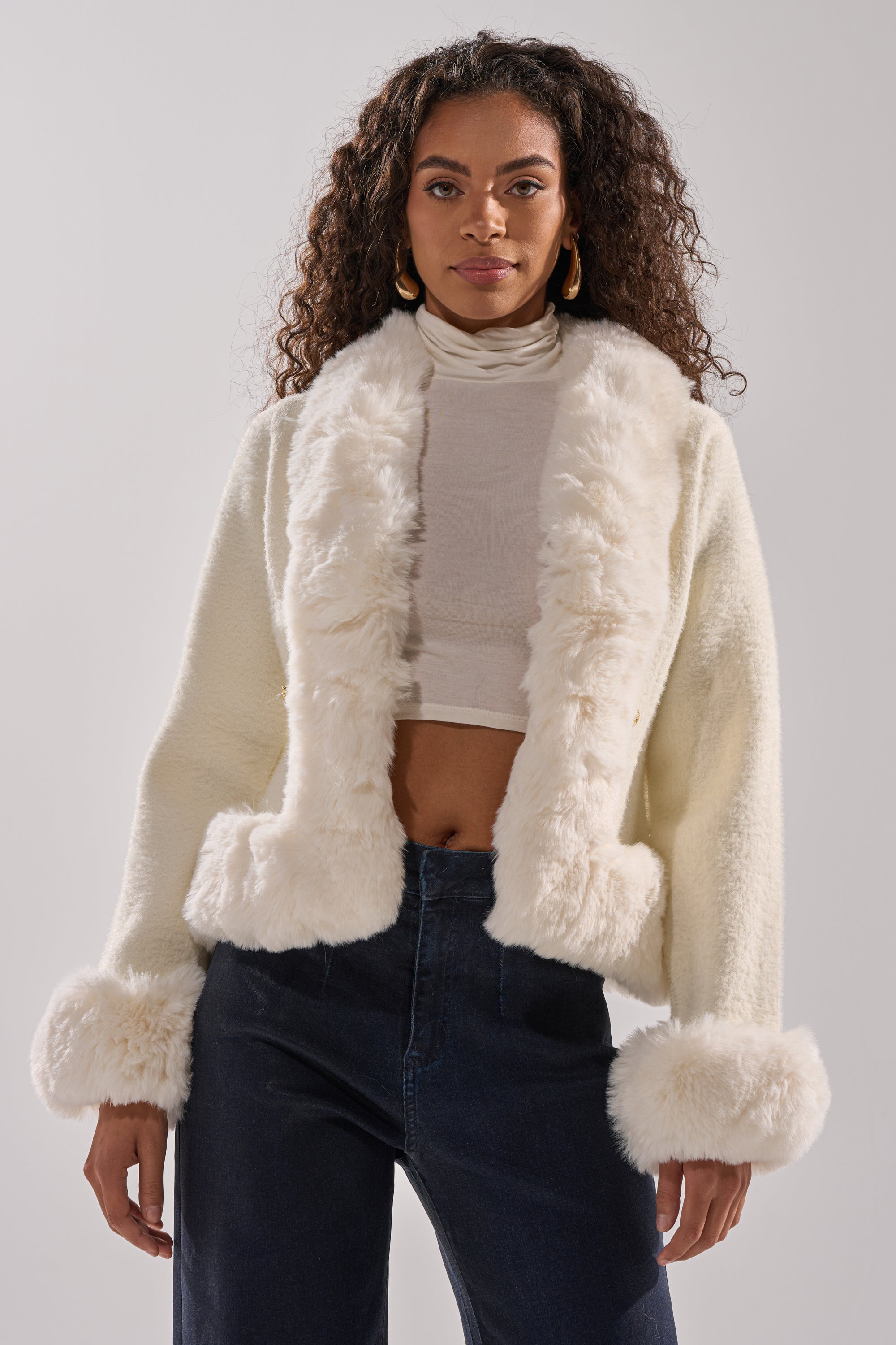 A woman with curly hair models the SOFT TOUCH CARDIGAN, featuring plush faux fur trim on the collar, cuffs, and hem, styled over a sheer white turtleneck crop top and dark high-waisted jeans against a plain background. 3