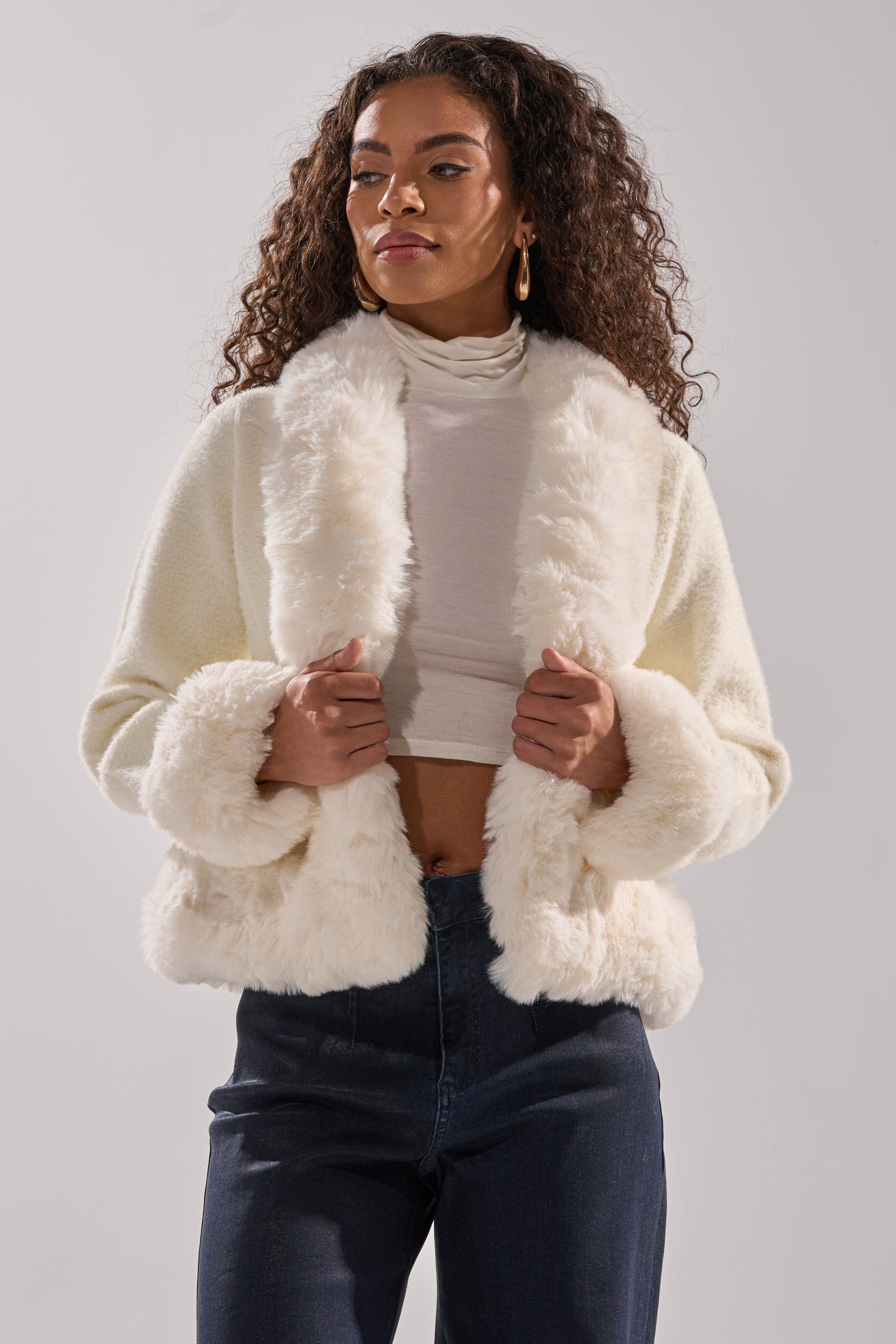 A woman with long curly hair wears the SOFT TOUCH CARDIGAN over a light turtleneck crop top and dark high-waisted jeans, standing confidently against a plain, light background. 4