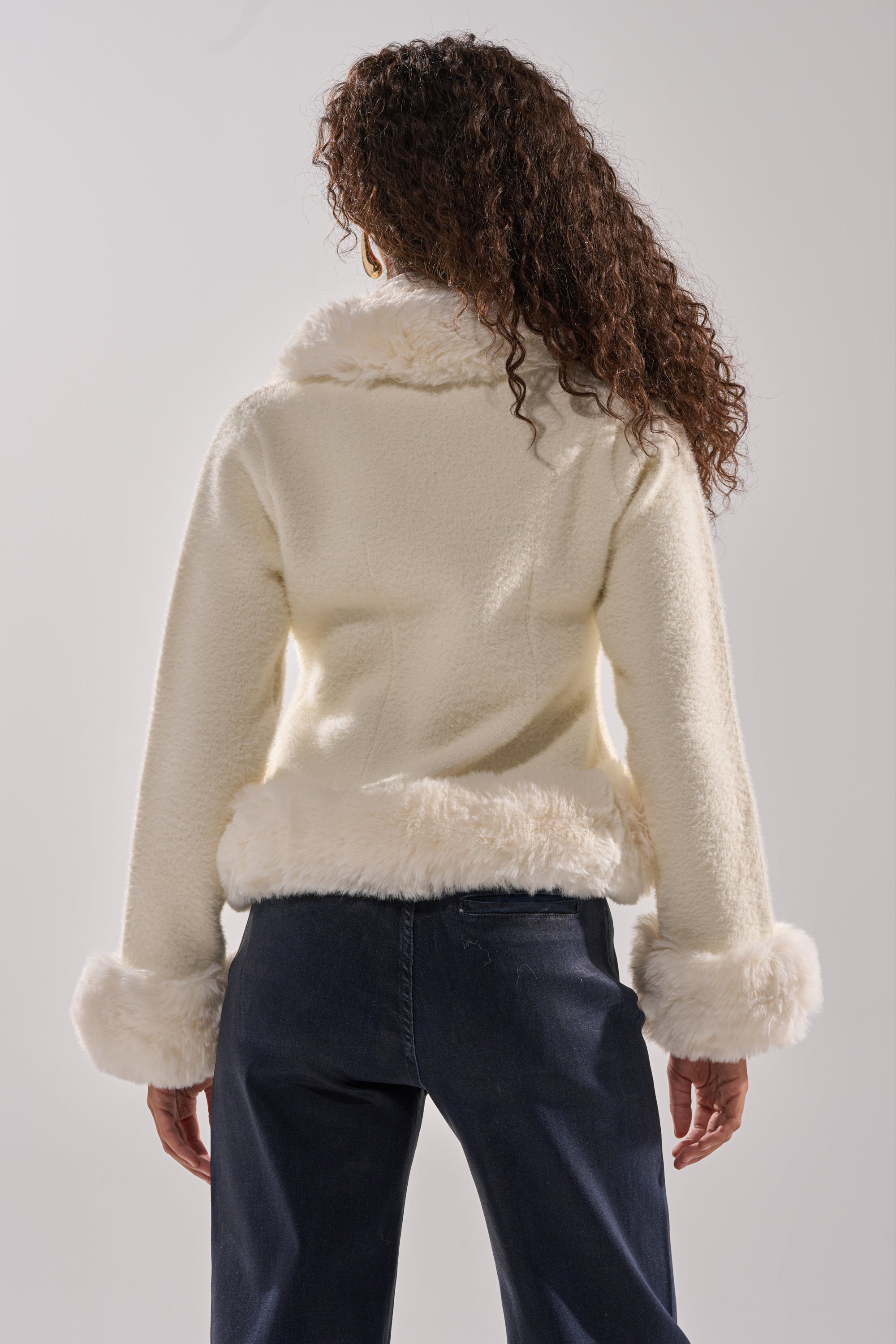 A person with long, curly hair is seen from the back wearing the SOFT TOUCH CARDIGAN, featuring cream faux fur trim with plush cuffs and hem, paired with dark jeans against a plain light background. 6