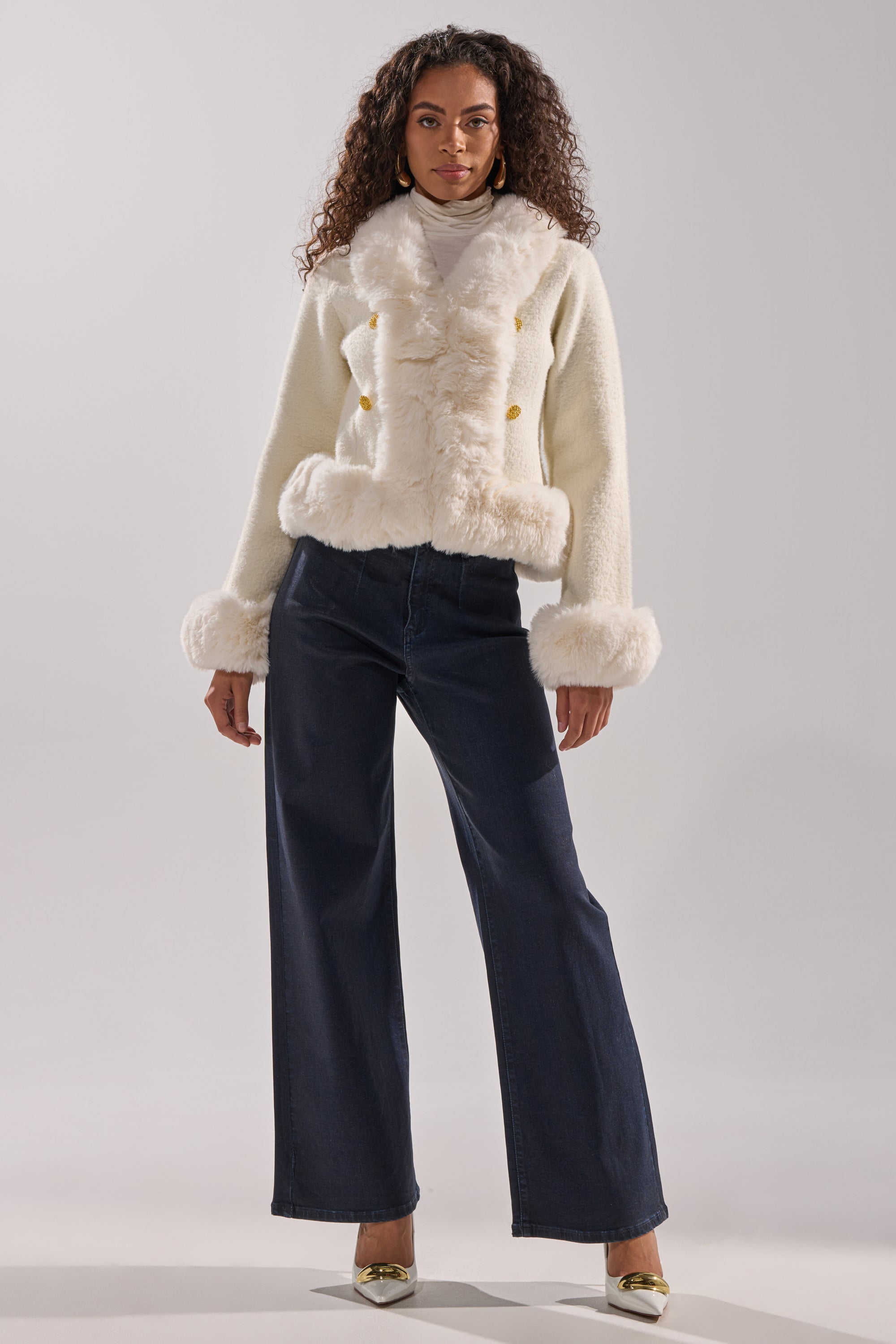 A woman stands facing forward wearing the SOFT TOUCH CARDIGAN with faux fur trim, wide-leg dark blue jeans, and metallic pointed-toe heels against a plain light gray background. 7