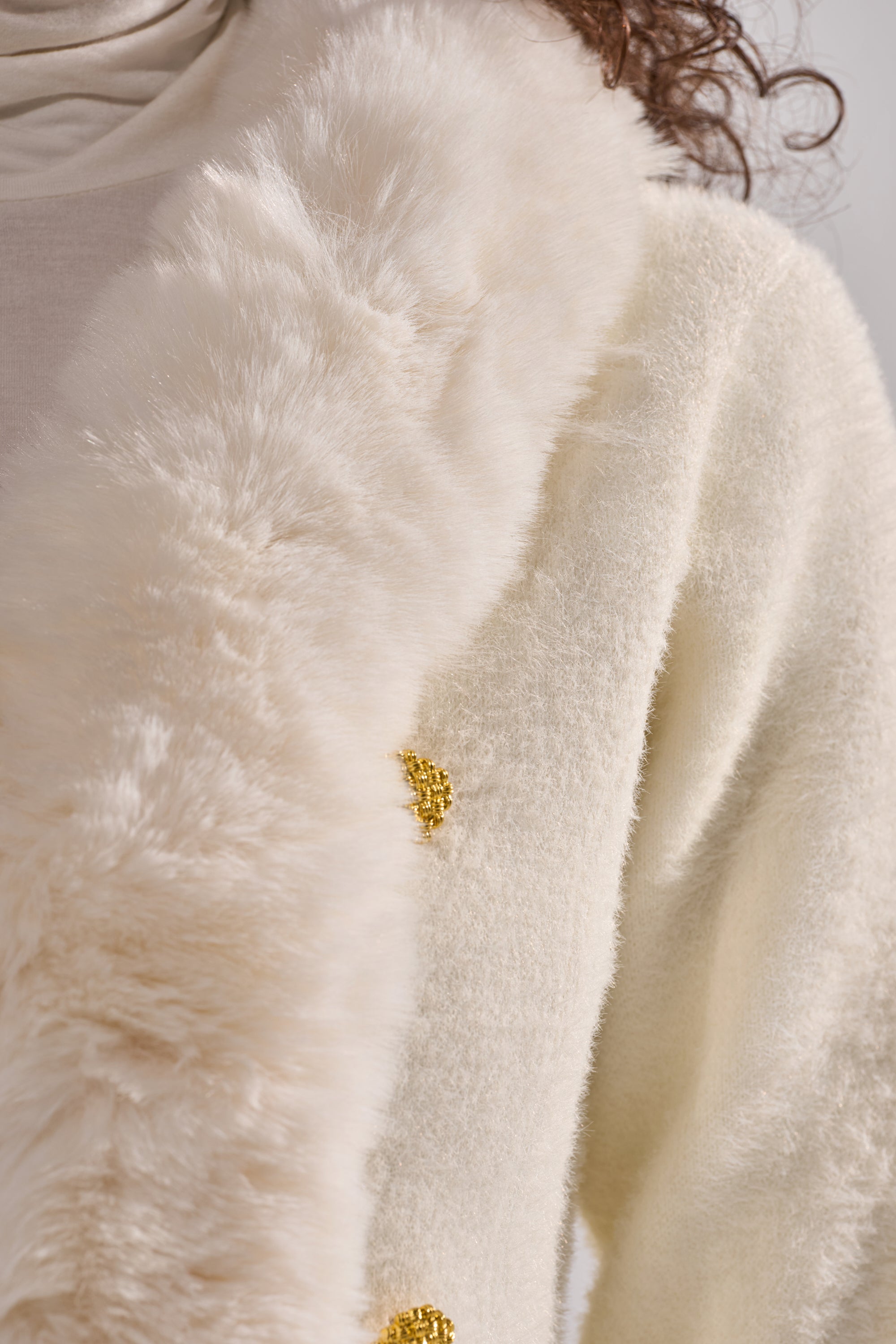 Close-up of a person in the SOFT TOUCH CARDIGAN in cream, showcasing its fuzzy texture, large faux fur collar, and gold decorative button. Only part of the figure is shown. 8