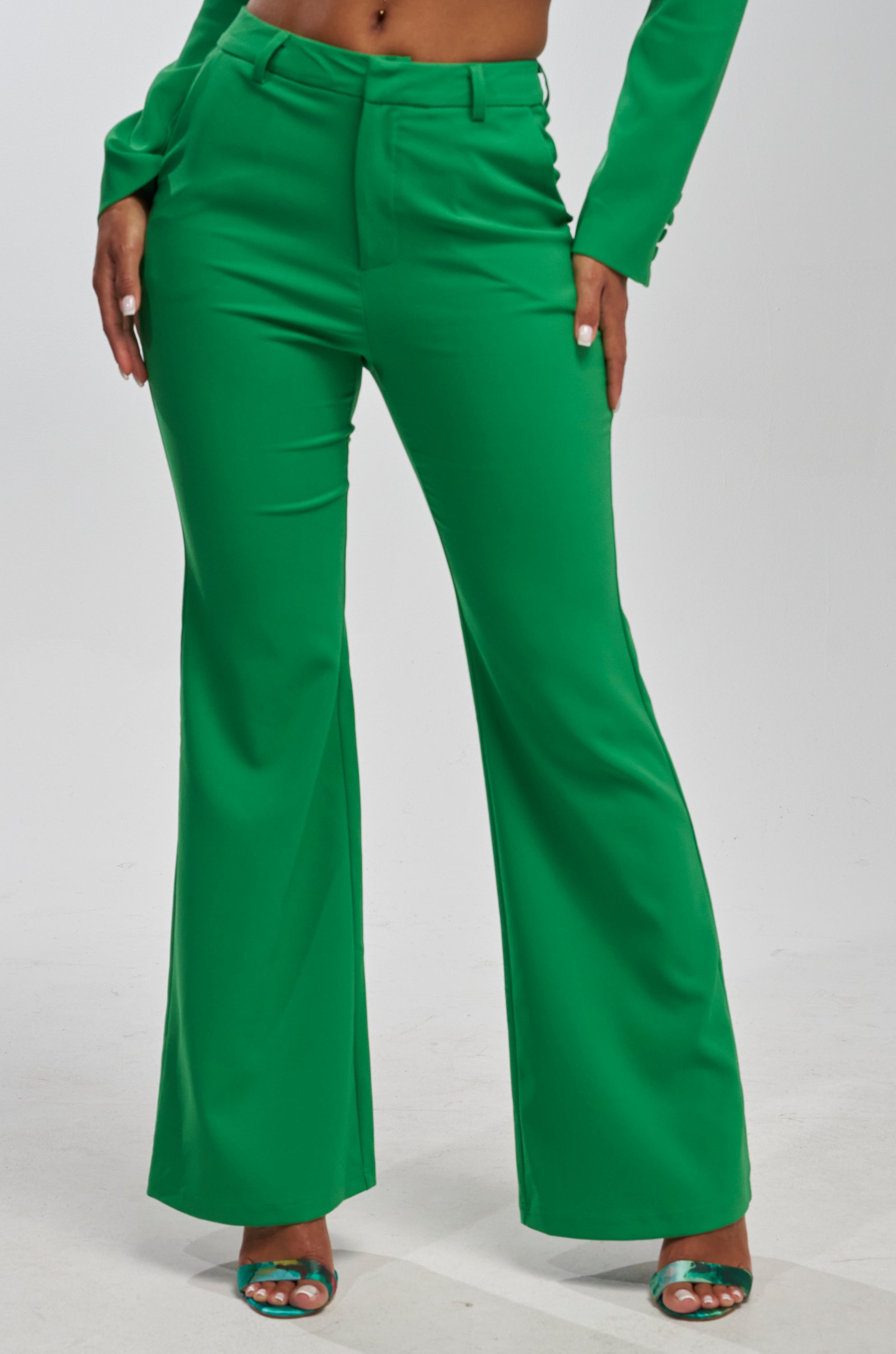 A woman wears the JUST YOUR LUCK FLARE TROUSER IN GREEN, styled with matching high-heeled sandals and a coordinating green blazer. The photo is cropped from the torso down.