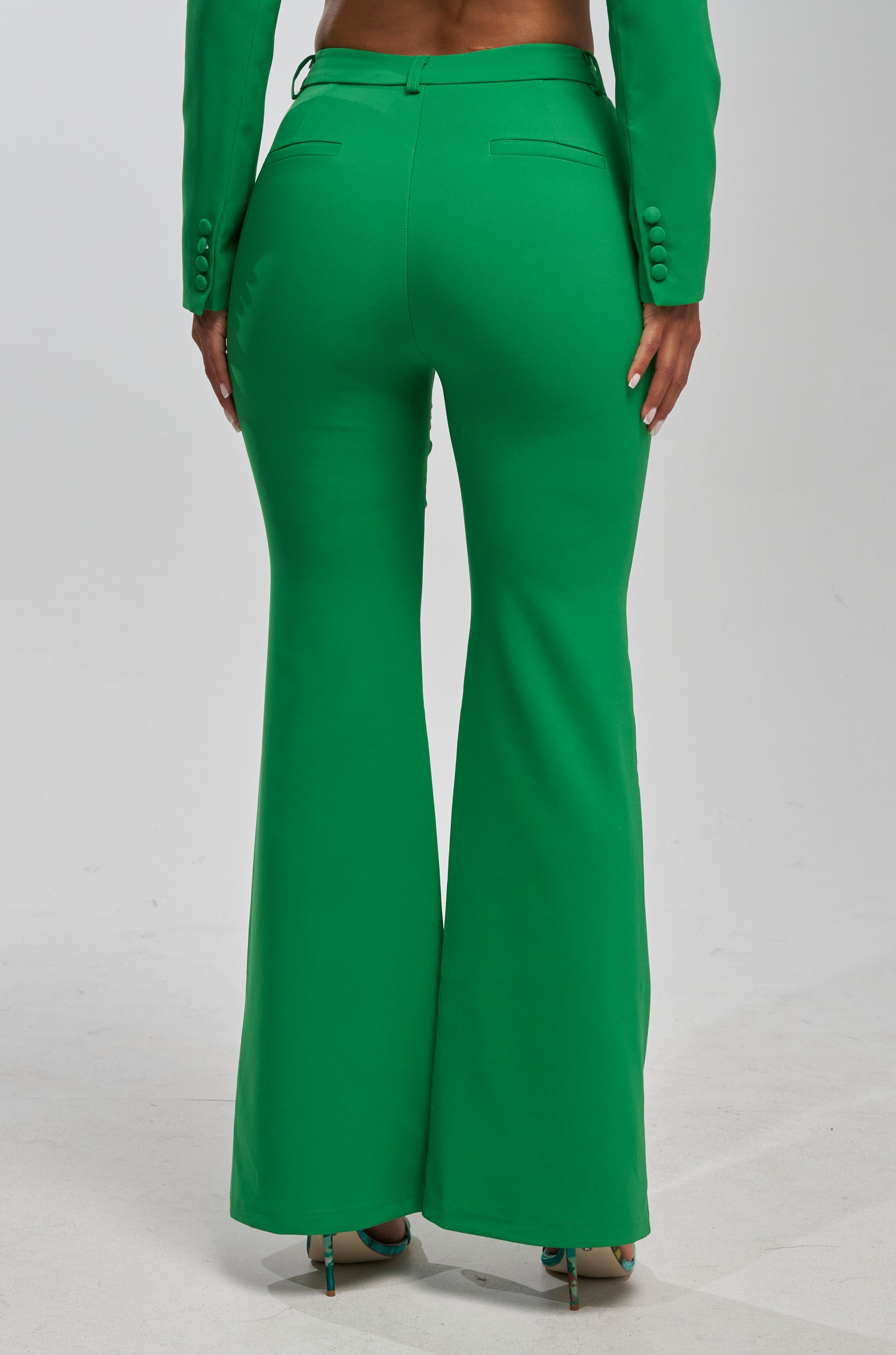 A person stands facing away from the camera on a light background, wearing the JUST YOUR LUCK FLARE TROUSER IN GREEN paired with a matching cropped blazer.