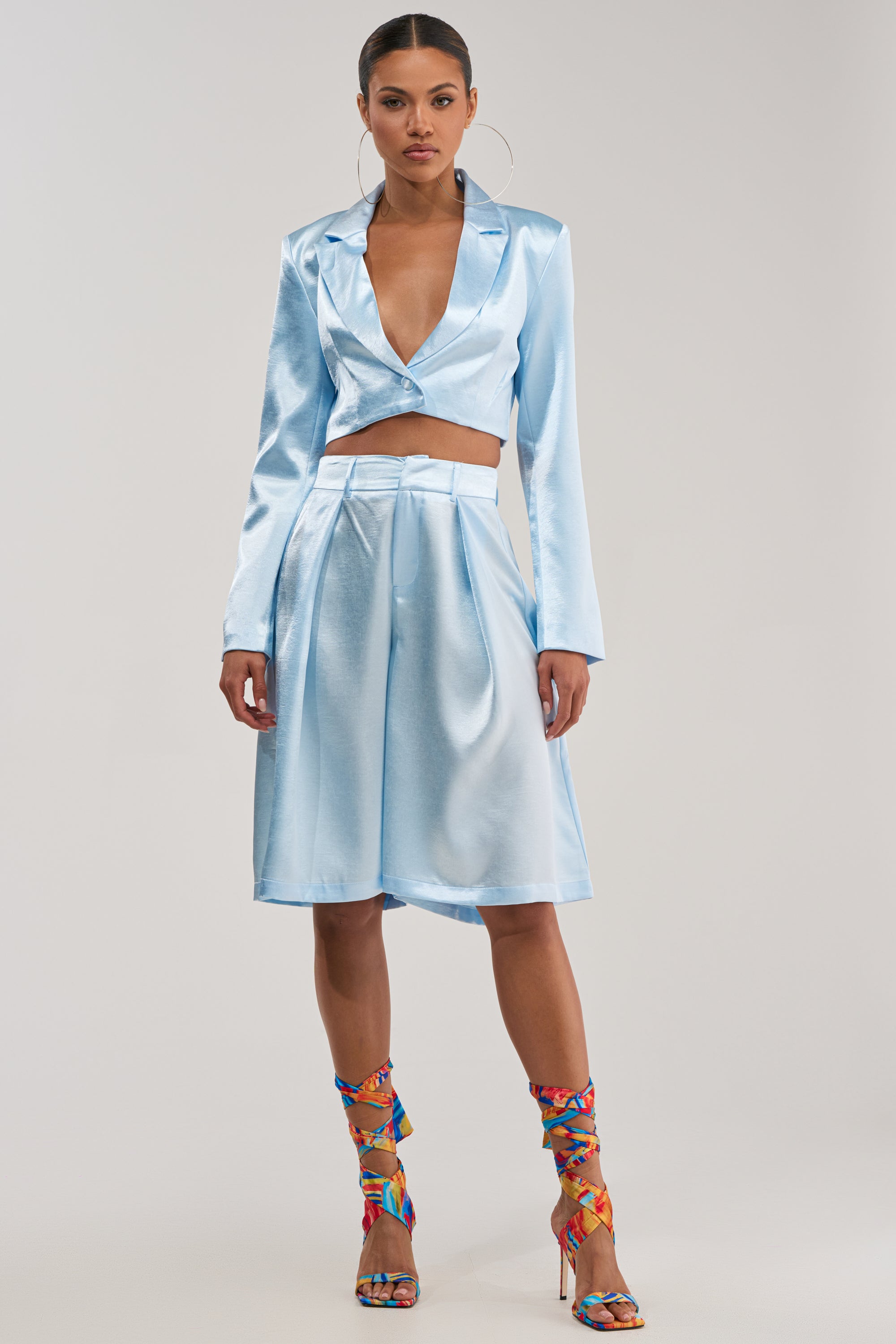 A woman poses in KYLA ICY BLUE SATIN TROUSER—featuring a sleek, modern fit—styled with a matching blazer, colorful strappy heels, and statement hoop earrings against a plain background for an elevated summer look.