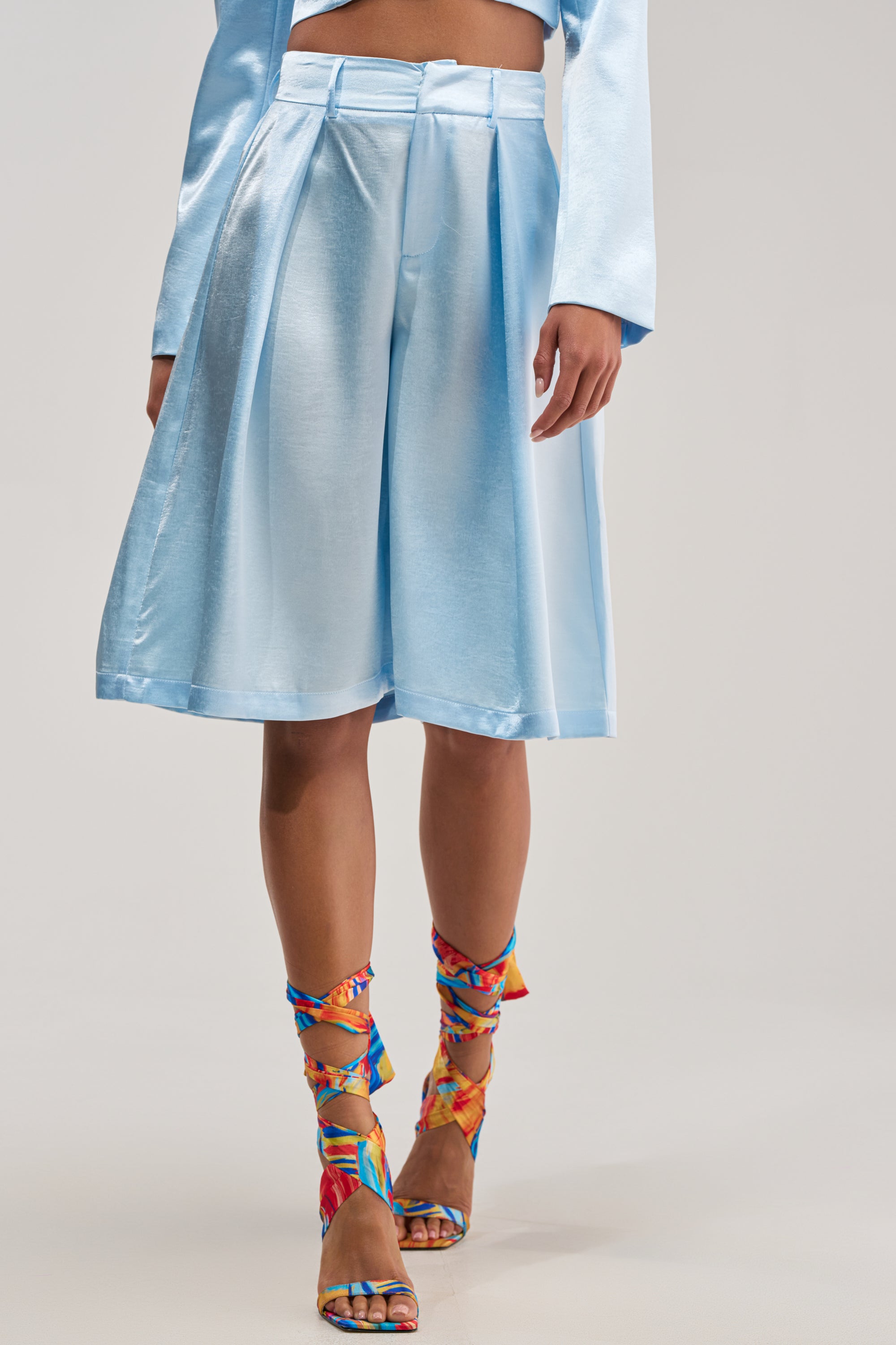 A person wearing the KYLA ICY BLUE SATIN TROUSER paired with a matching long-sleeve top and colorful, strappy high-heeled sandals—ideal for a chic summer date night against a plain background.