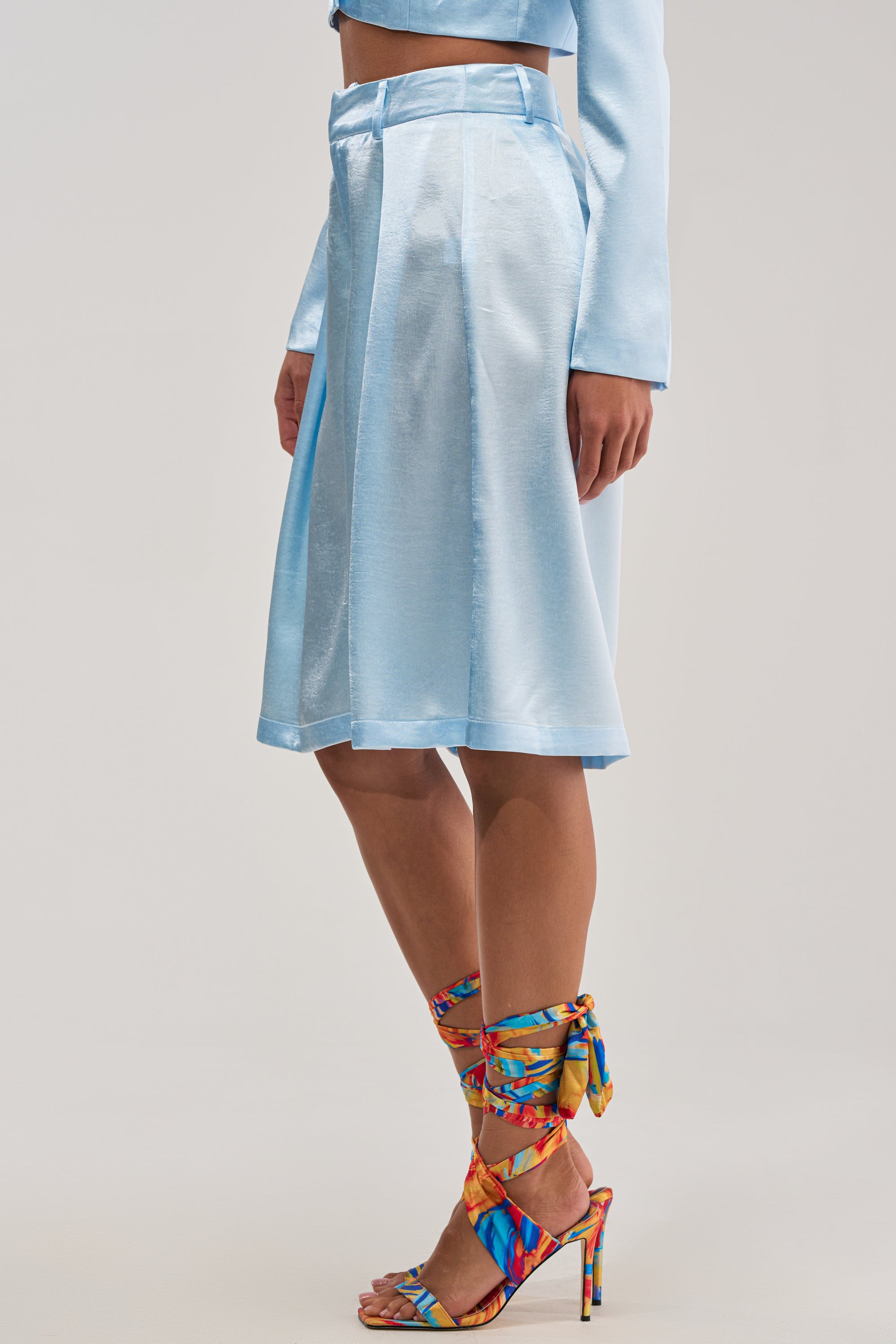 A person wearing the KYLA ICY BLUE SATIN TROUSER with a matching crop top and vibrant ankle-tie heels stands against a neutral background—an effortlessly chic look for summer date nights.