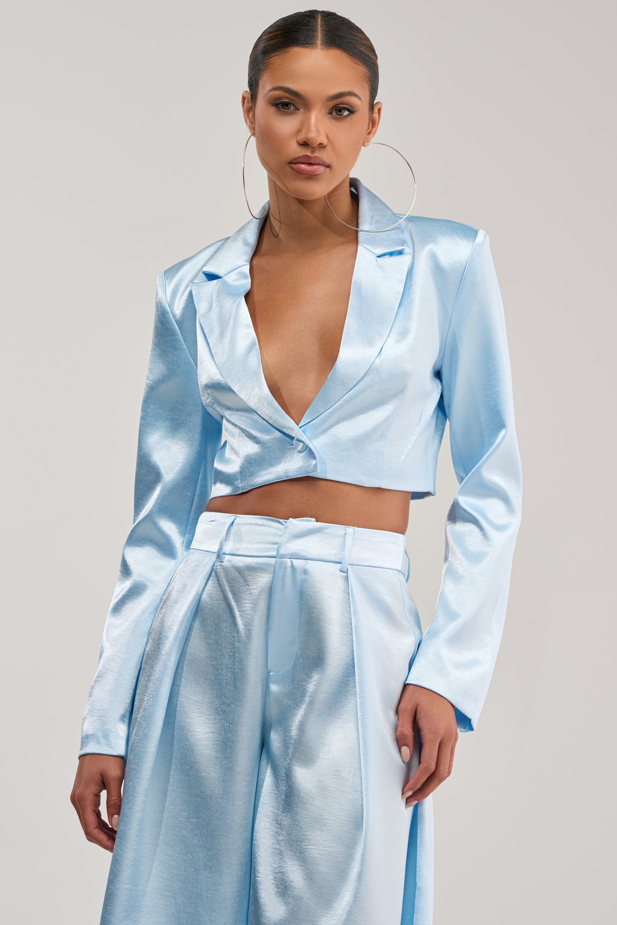 A woman poses confidently against a light gray background in the KYLA ICY BLUE SATIN BLAZER paired with wide-leg pants—a chic and elegant choice for a summer date night.
