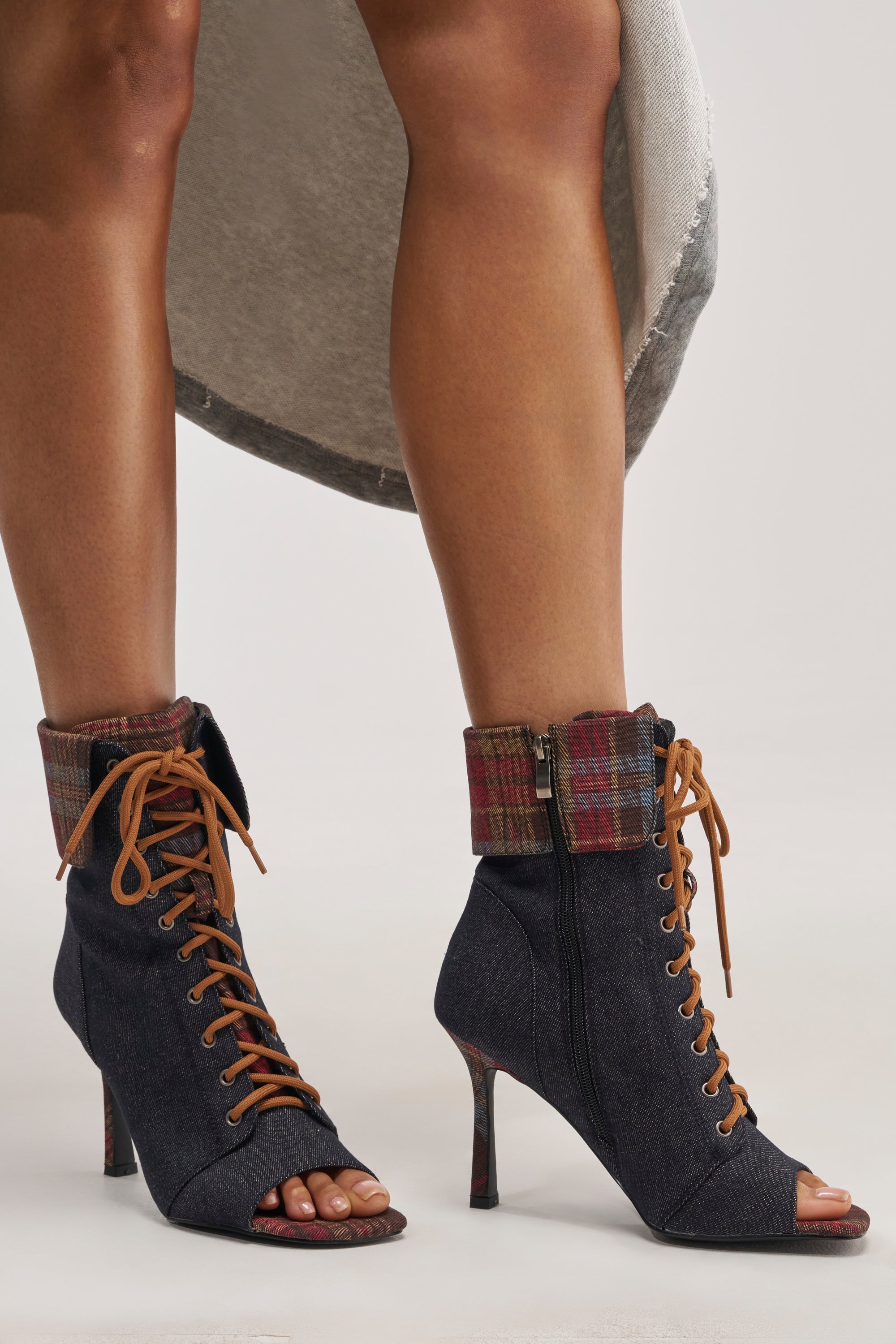 Wearing the AZALEA WANG KYLAN DENIM PATCHWORK OPEN TOE BOOTIE—high-heeled lace-up boots in dark denim with brown laces, plaid cuffs, side zippers, and bold plaid detailing on the heels—a person shows off a glimpse of their skirt above these unique streetwear booties.