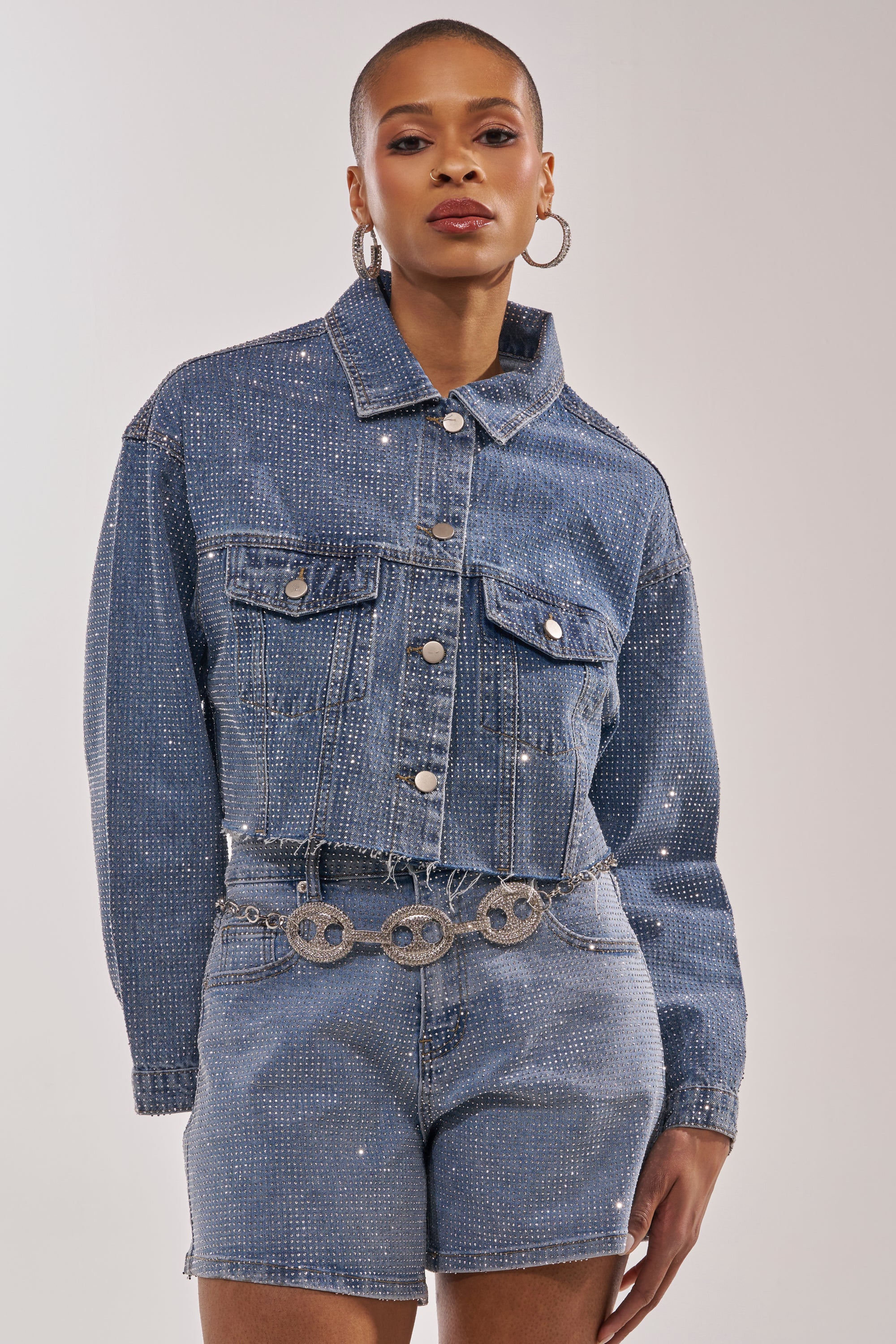 A woman poses confidently in the KYLIE EMBELLISHED DENIM JACKET and matching high-waisted shorts, styled with hoop earrings and a silver chain belt featuring circular details. 4