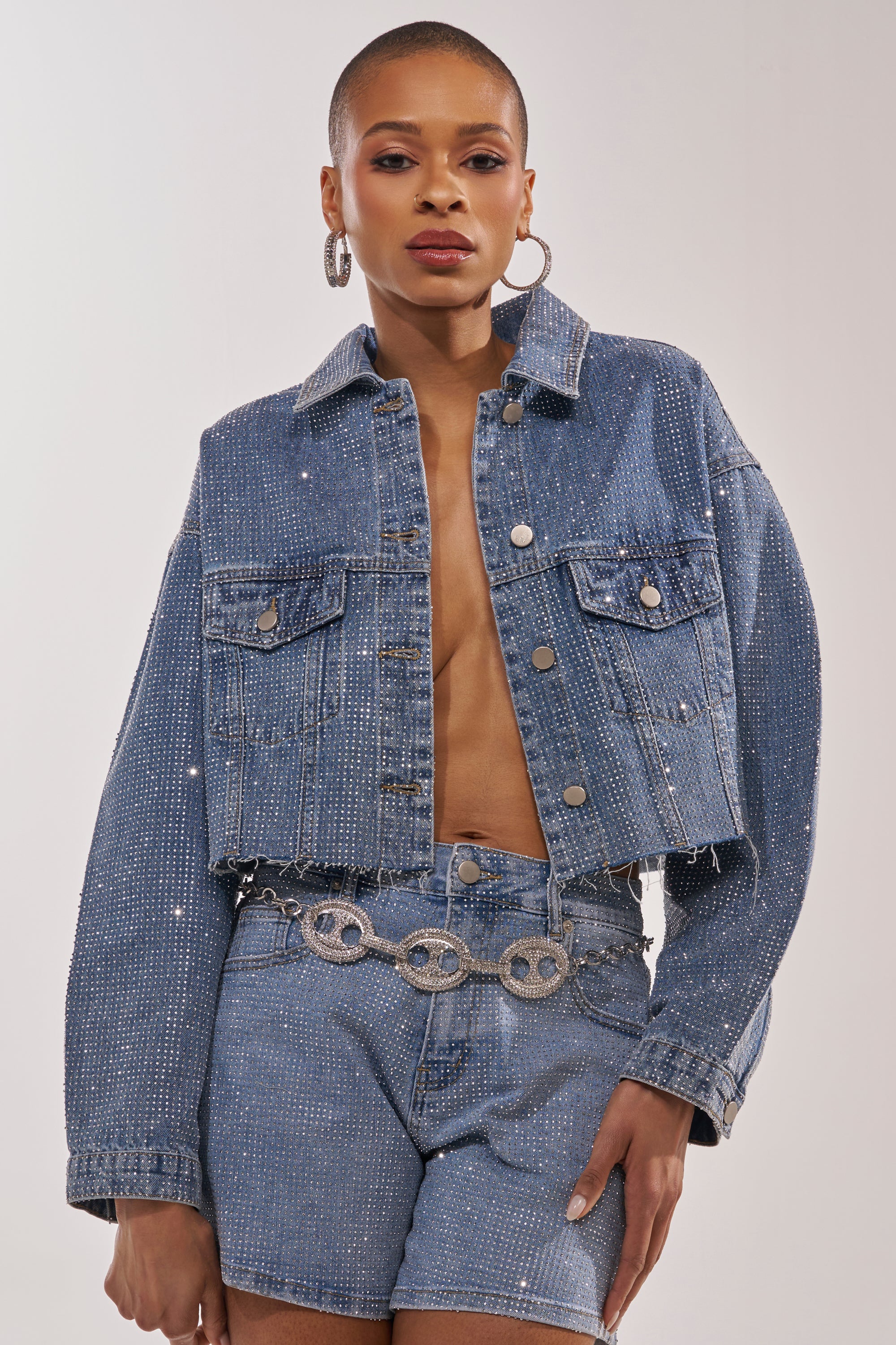 A stylish person wears the KYLIE EMBELLISHED DENIM JACKET with matching high-waisted shorts, both adorned with sparkling details. Hoop earrings and a silver chain belt complete the look. 5