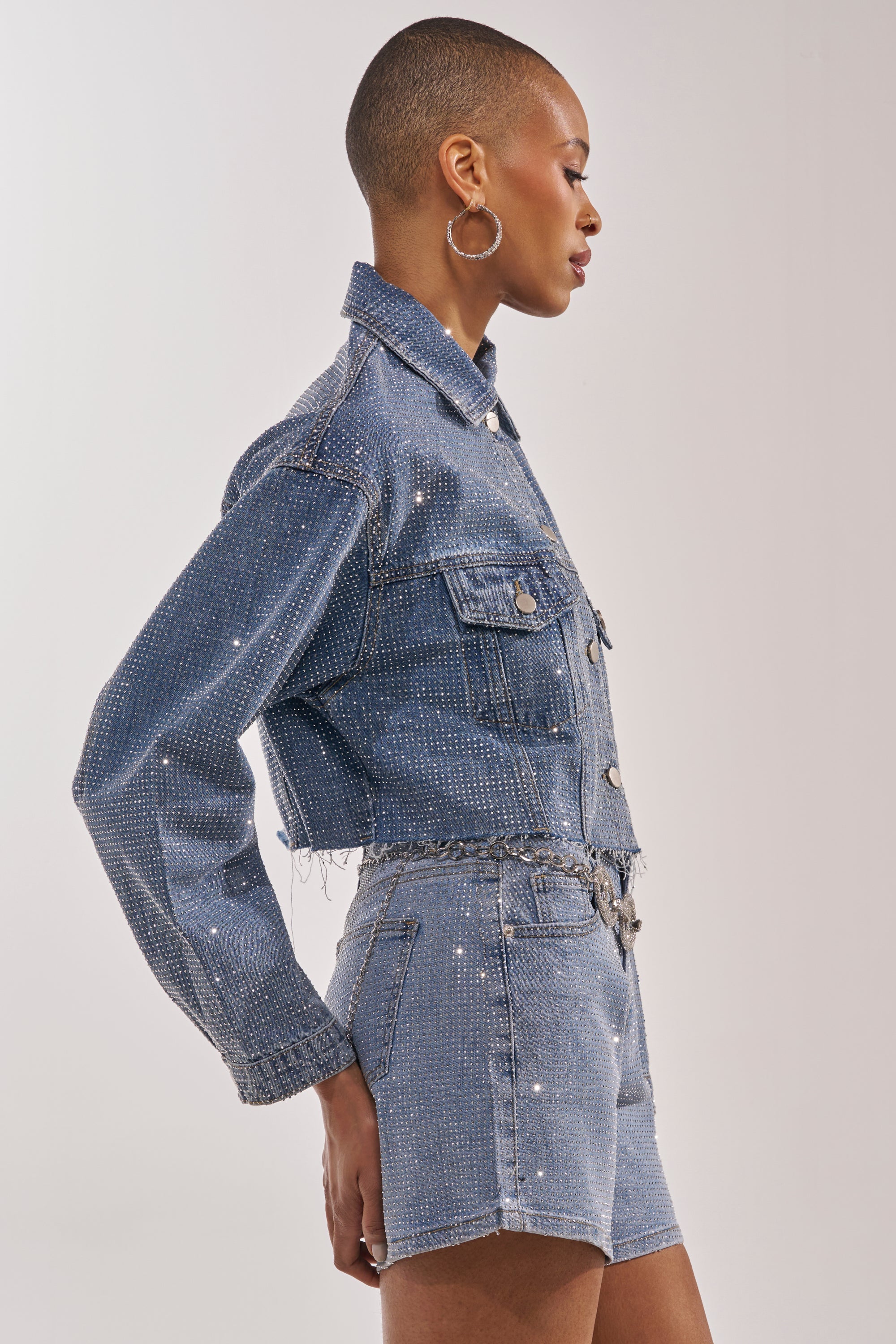 A person with short hair models the KYLIE EMBELLISHED DENIM JACKET, styled with matching high-waisted shorts and sparkling details. They pose confidently sideways, wearing large hoop earrings against a plain background. 6