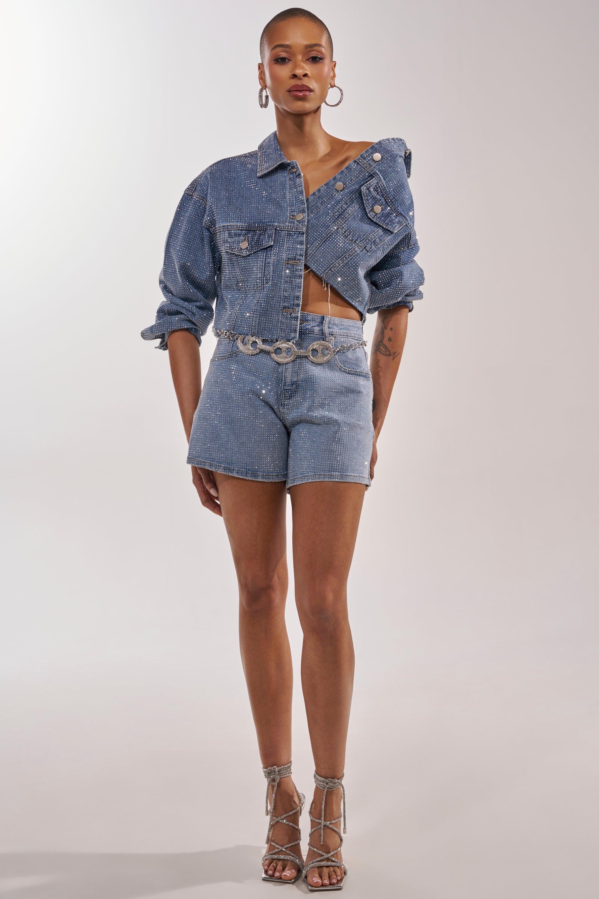 Wearing the KYLIE EMBELLISHED DENIM JACKET, high-waisted denim shorts, a chain belt with circular links, hoop earrings, and strappy heels, a person stands against a plain light background. 9