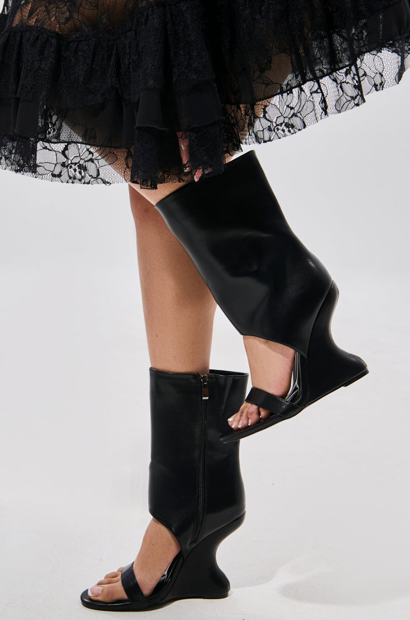 A person pairs a black lace skirt with the AZALEA WANG KYVEKI BLACK WEDGE OPEN TOE BOOTIE, featuring clear straps and curved wedge soles for a bold, edgy touch.