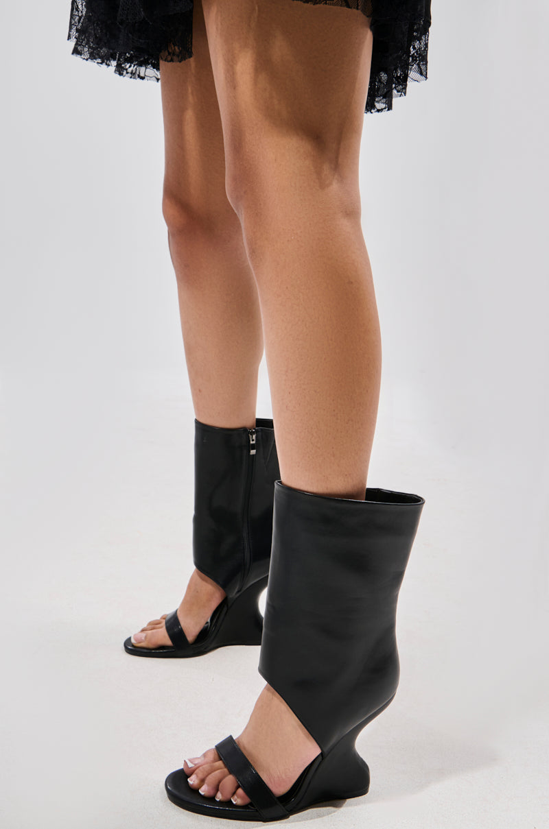 A person wears AZALEA WANG KYVEKI BLACK WEDGE OPEN TOE BOOTIE heels with a sculpted design, standing on a light surface. The hem of a black lace skirt appears above—an edgy twist on faux leather booties.