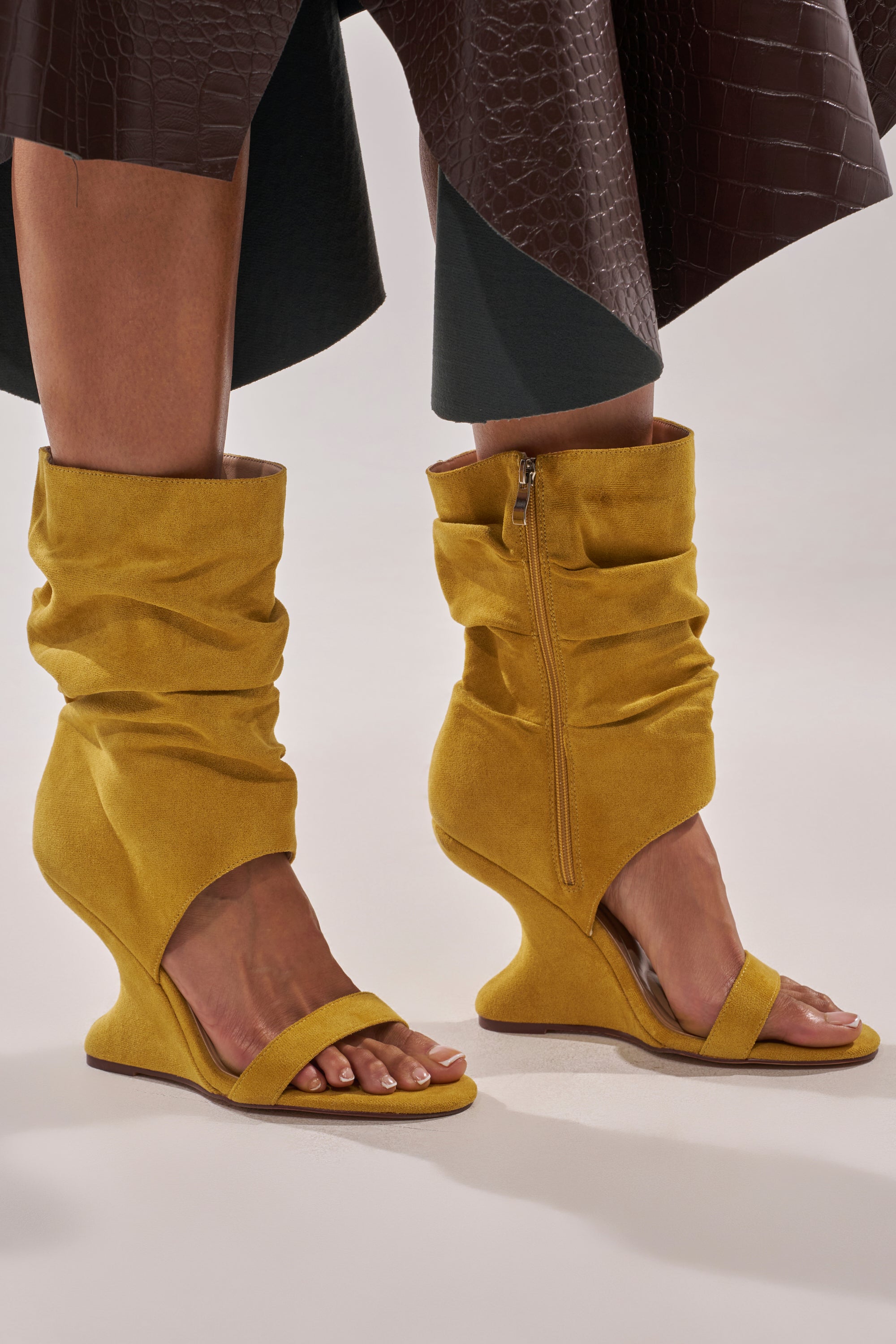 A person wears the AZALEA WANG KYVELISA MUSTARD WEDGE OPEN TOE BOOTIE in mustard yellow faux suede, featuring sculptural wedge heels, open toe design, and zipper sides, while standing on a light surface. 1