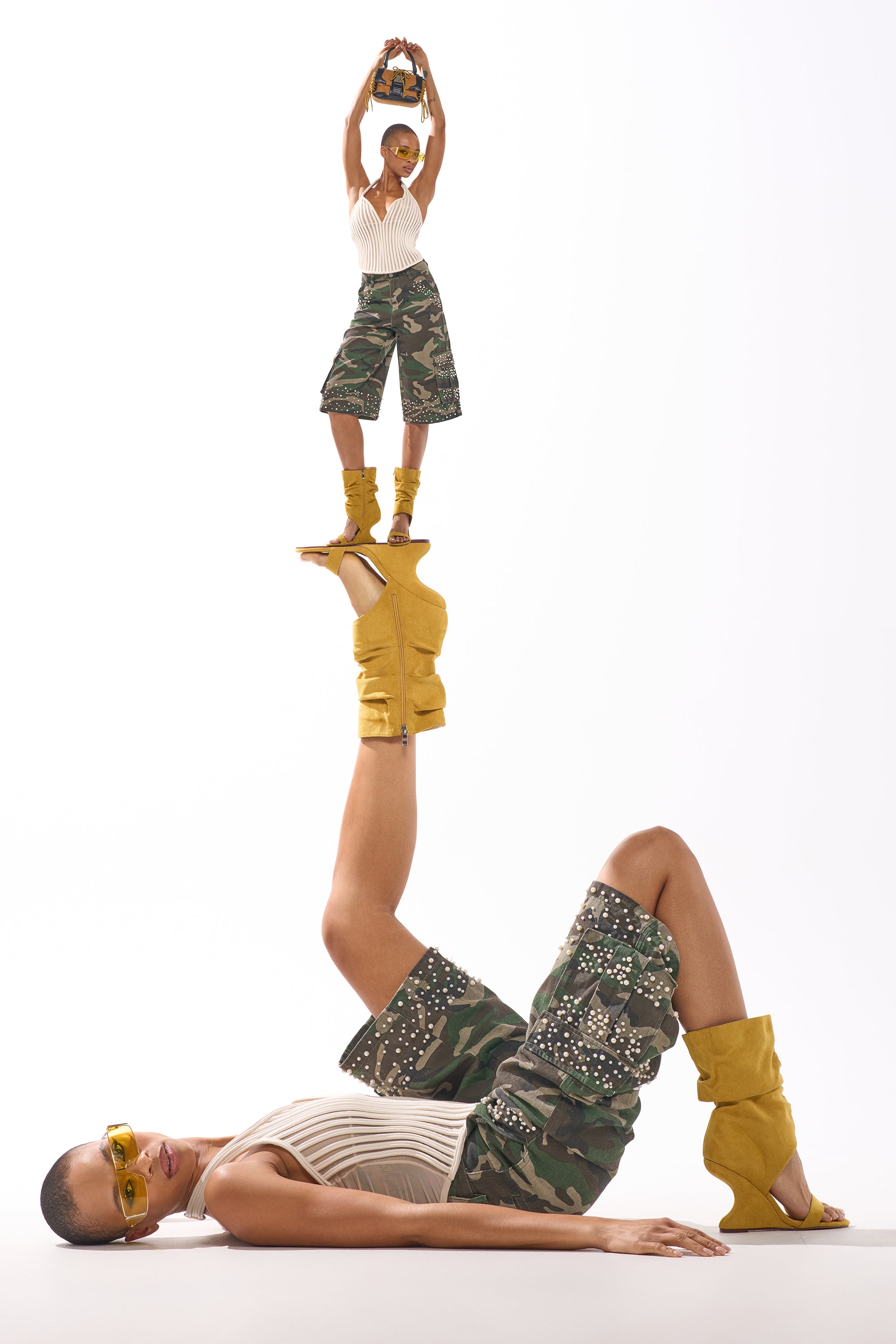 A woman in camo shorts, a white top, and AZALEA WANG KYVELISA MUSTARD WEDGE OPEN TOE BOOTIE lies on her back with one leg raised. A miniature version of herself stands balanced on her foot holding a small purse overhead. 0