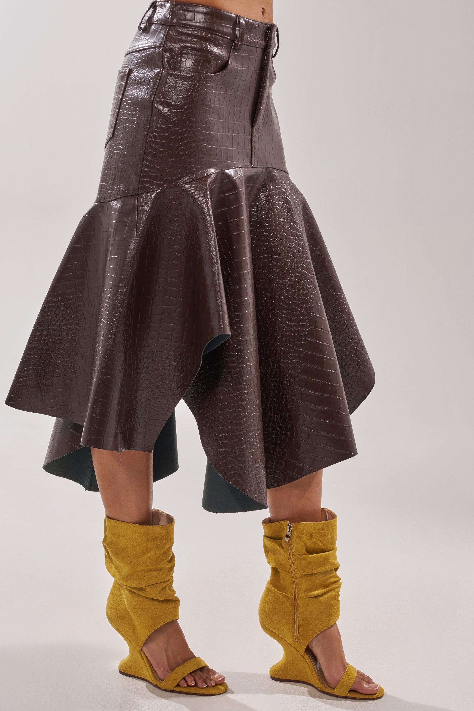 A person wears a dark brown, crocodile-textured flared midi skirt with AZALEA WANG KYVELISA Mustard Wedge Open Toe Booties featuring ruched details and zippers, set against a plain light gray background. 2