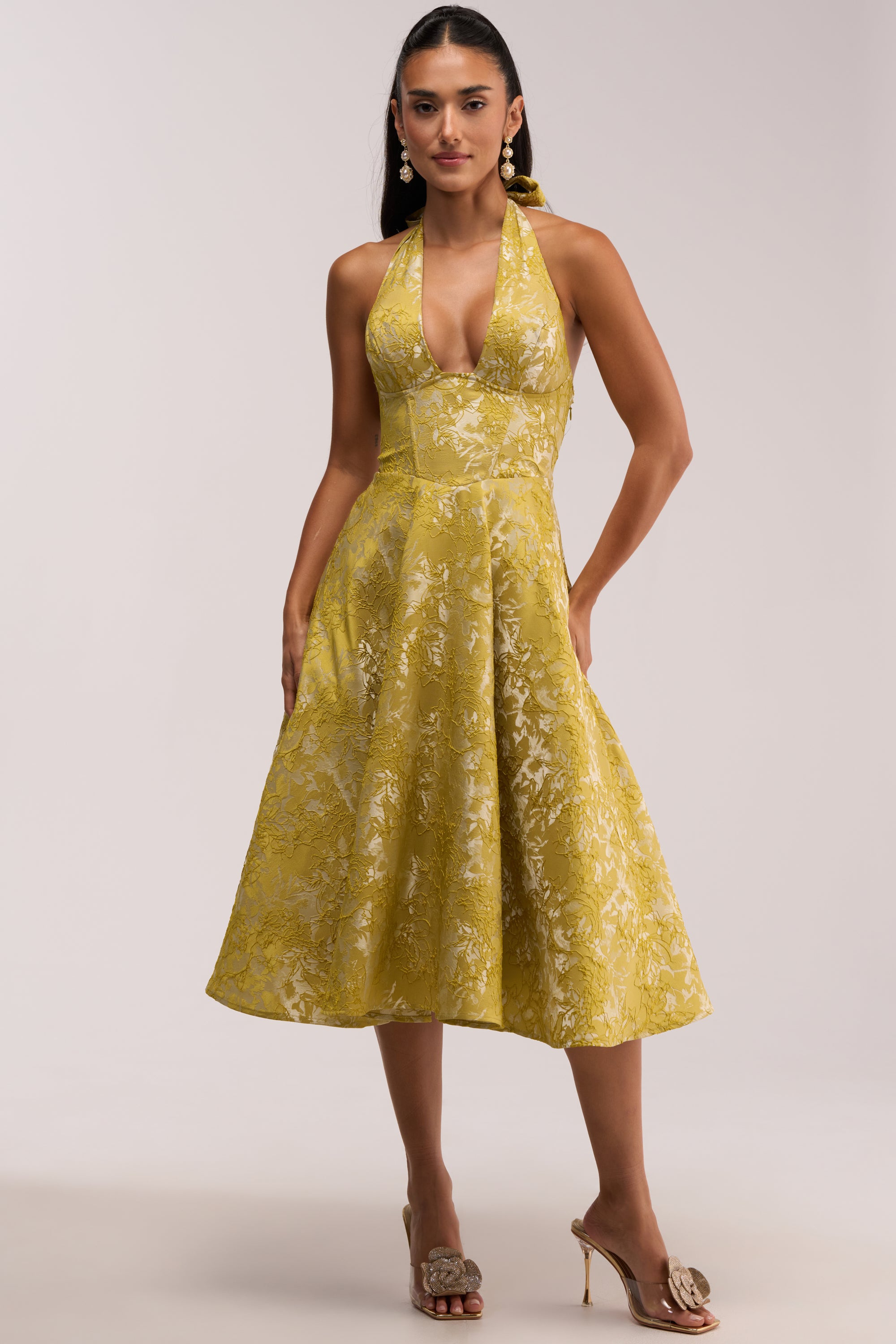 A woman wearing the SWEET CAROLINE HALTER MIDI DRESS IN GREEN, featuring a vibrant green floral brocade fabric, halter neckline, deep V-cut, and full skirt reminiscent of an embroidered summer dress, paired with nude heeled sandals and pearl drop earrings.
