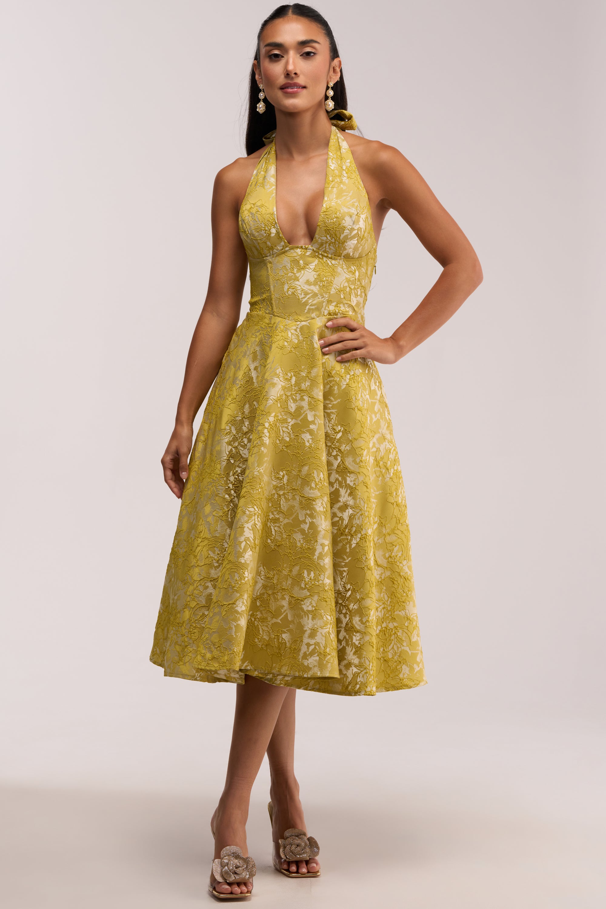 A woman wearing the SWEET CAROLINE HALTER MIDI DRESS IN GREEN with open-toe shoes. She has long hair, statement earrings, and is standing against a plain light background.
