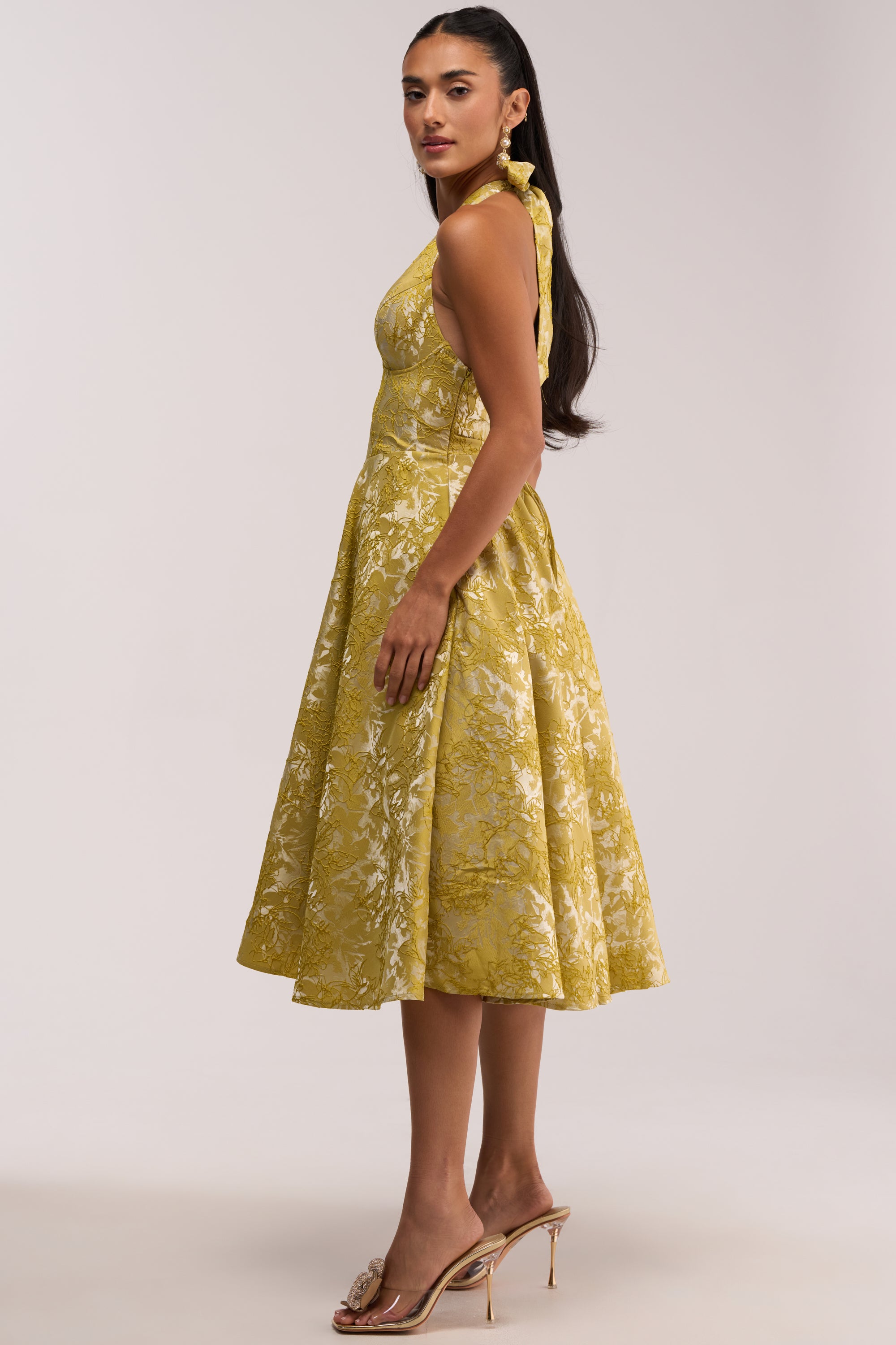 A woman stands sideways against a plain light-colored background, wearing the SWEET CAROLINE HALTER MIDI DRESS IN GREEN—a sleeveless halter midi dress with a floral pattern. She has long dark hair styled in a ponytail, accessorized with gold earrings and heeled sandals.