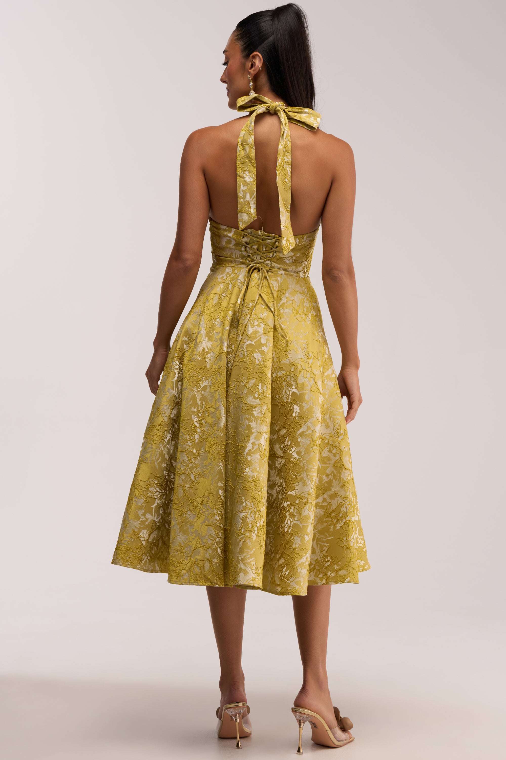 A woman stands facing away, wearing the SWEET CAROLINE HALTER MIDI DRESS IN GREEN, which features a halter neckline with a tied bow, a fitted waist, and a flared skirt. She has long dark hair in a ponytail and is paired with high-heeled sandals.