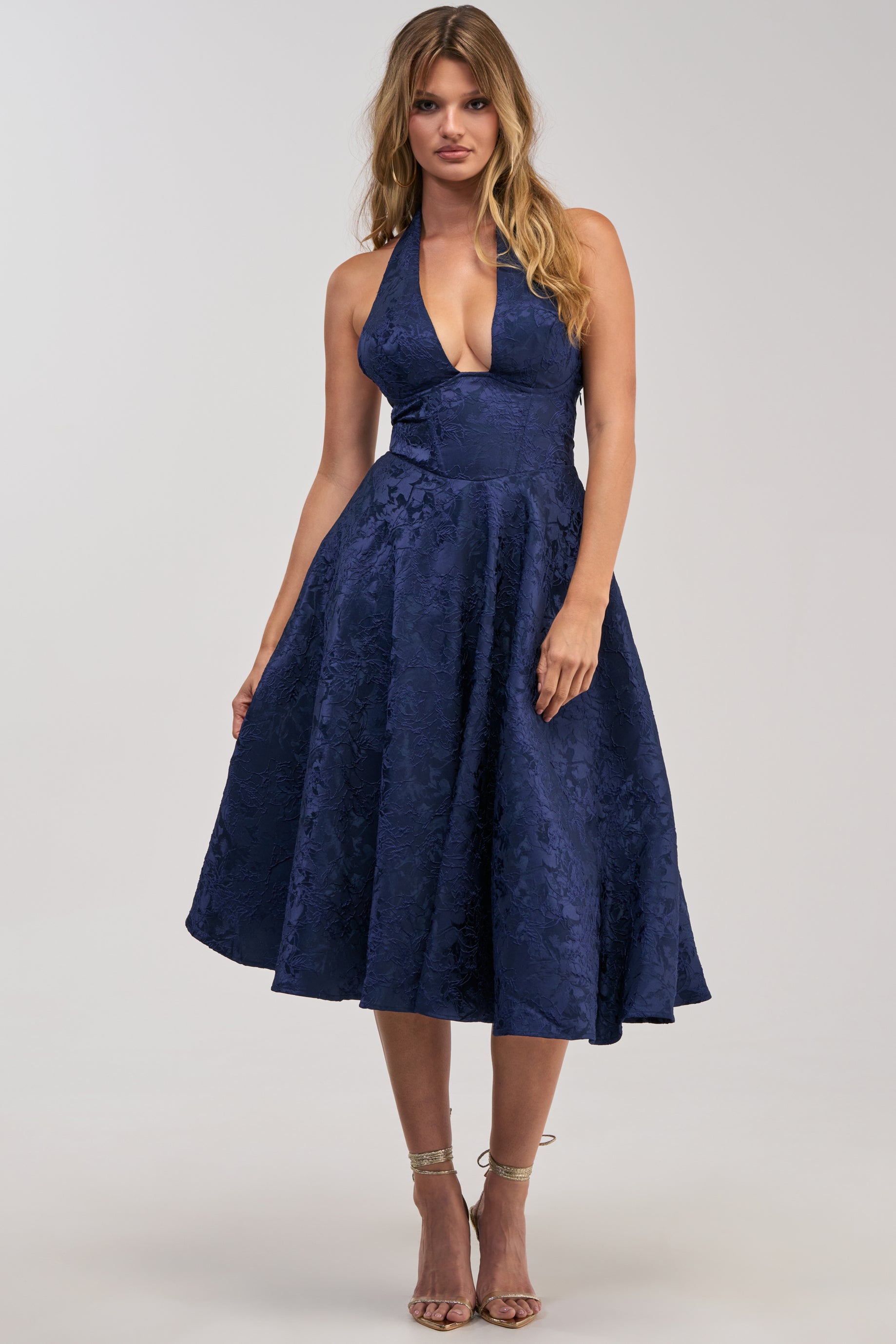 A woman wearing the SWEET CAROLINE HALTER MIDI DRESS IN NAVY—a sleeveless, halter-neck, knee-length cocktail party dress with a flared skirt and subtle floral texture—paired with strappy high-heeled sandals.
