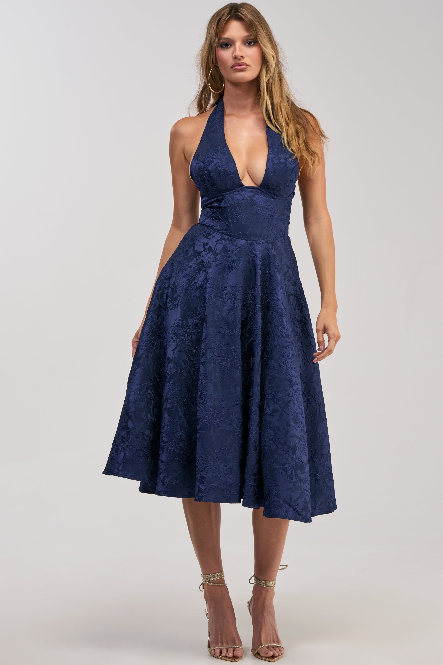 A woman wearing the SWEET CAROLINE HALTER MIDI DRESS IN NAVY—a sleeveless, halter-neck, navy blue midi dress with a flared skirt and floral pattern—paired with strappy high-heeled sandals, stands against a plain background.