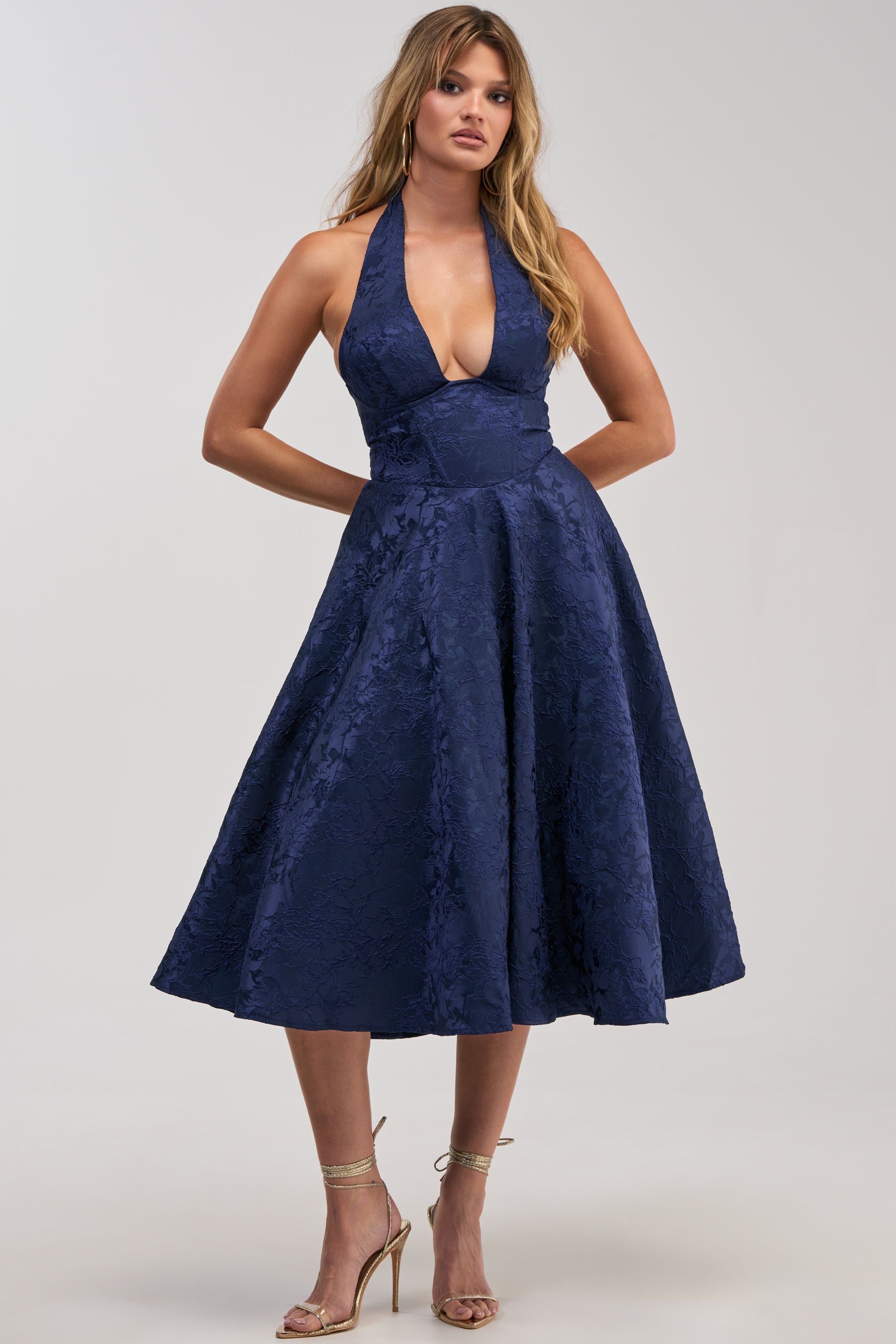 Dressed in the SWEET CAROLINE HALTER MIDI DRESS IN NAVY—a sleeveless, deep V-neck halter midi featuring a flared skirt and floral textured fabric—she pairs it with strappy high-heeled sandals as she stands against a neutral background, making this the ideal choice for an elegant evening or any cocktail party.