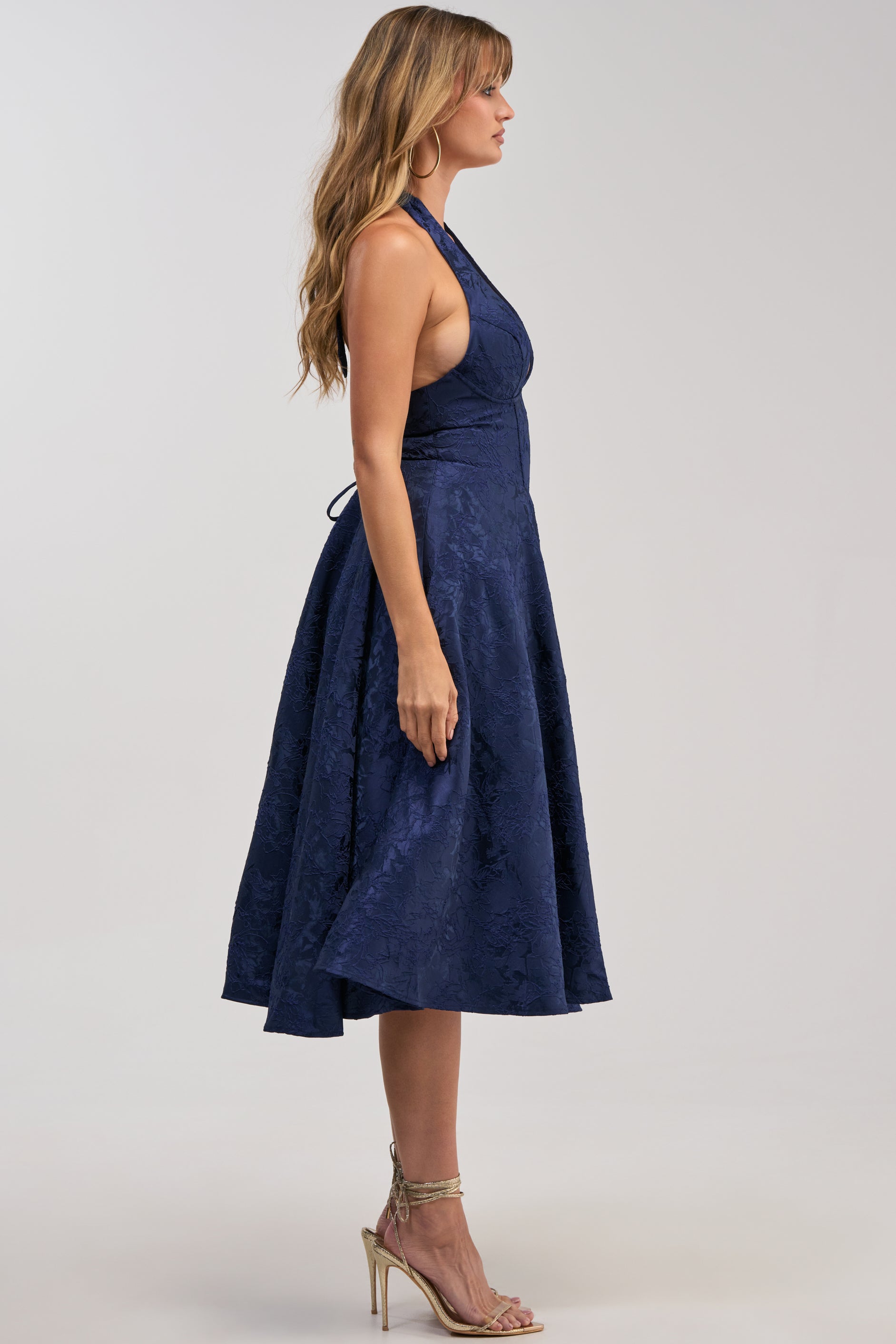 Wearing the SWEET CAROLINE HALTER MIDI DRESS IN NAVY, a woman stands in profile with long wavy hair, hoop earrings, and strappy high-heeled sandals—the perfect sleeveless, flared look for any cocktail party or evening event.