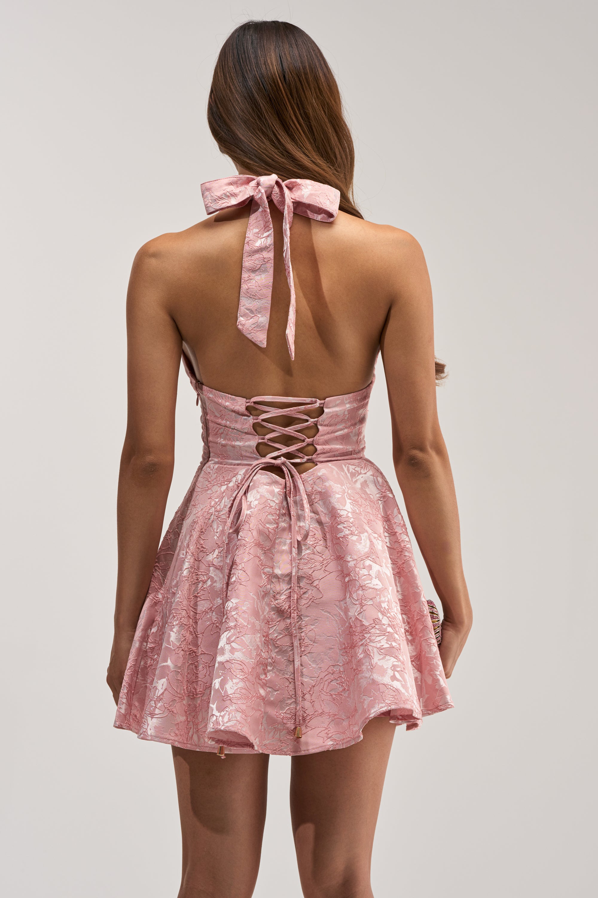 A woman is seen from behind wearing the TALK IS CHEAP FLORAL MINI DRESS—a shiny pink, floral-patterned halter mini dress featuring a lace-up back and flared skirt.