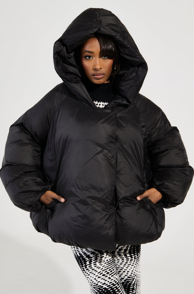 A person stands confidently, hands in pockets, wearing a large black Leone Statement Circle Crop Puffer with a hood. Their patterned black and white pants perfectly complement the bold jacket. The plain, neutral background highlights their striking style.