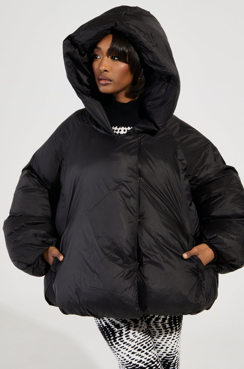The person is confidently wearing a LEONE STATEMENT CIRCLE CROP PUFFER IN BLACK, with hands casually in pockets, paired with black and white patterned pants. They gaze to the side against a plain background, exuding effortless style.