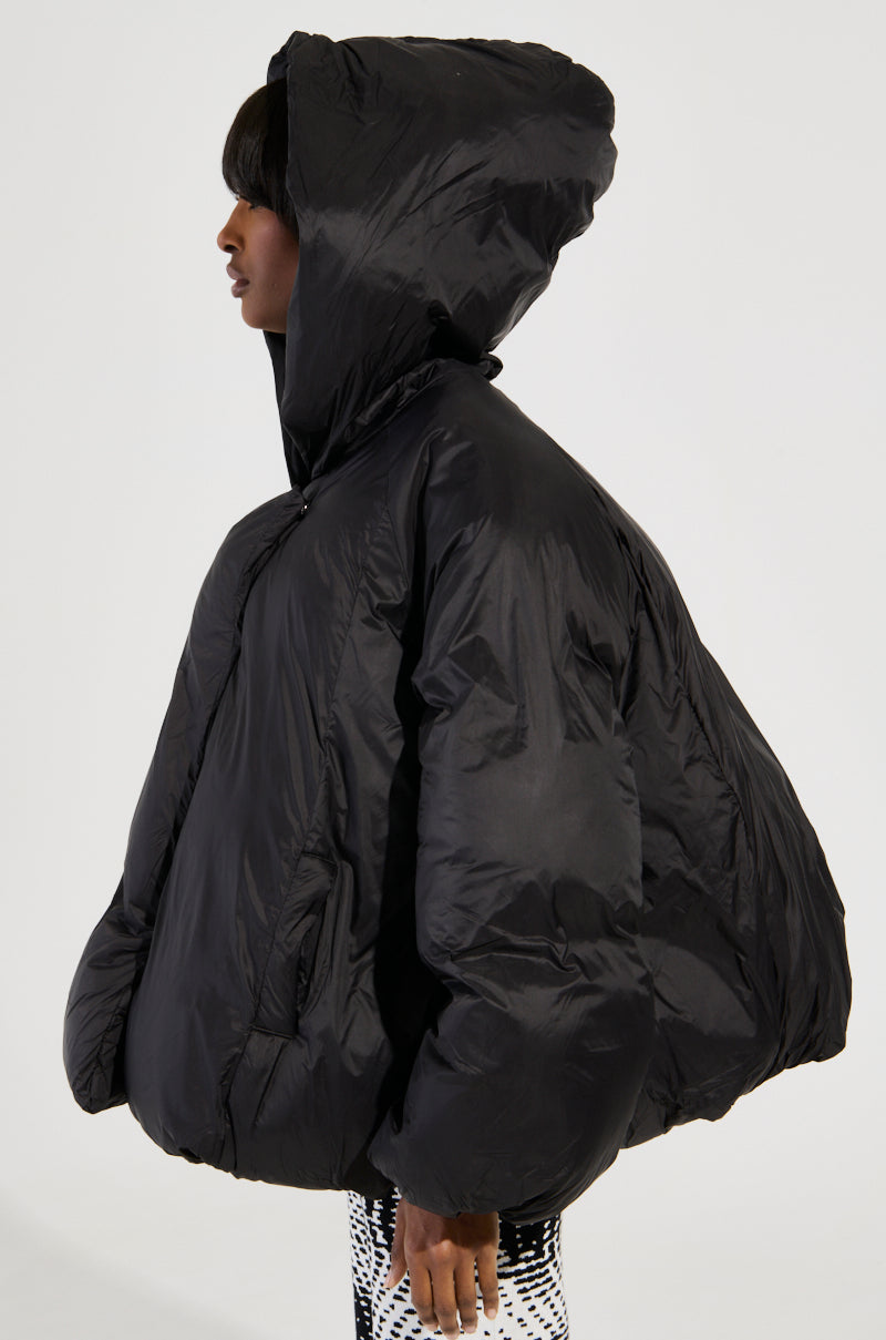 A person is shown in profile wearing the LEONE STATEMENT CIRCLE CROP PUFFER IN BLACK, a stylish oversized hooded jacket featuring a voluminous design and A-line hem, complemented by patterned pants. The jacket completely covers the person's upper body against a plain white background.