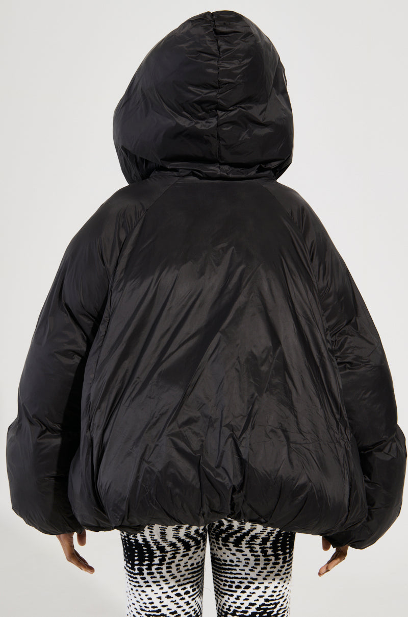 A person wearing the Leone Statement Circle Crop Puffer in black with double-breasted closures stands facing away. They also wear black and white patterned pants. The background is plain and light gray.