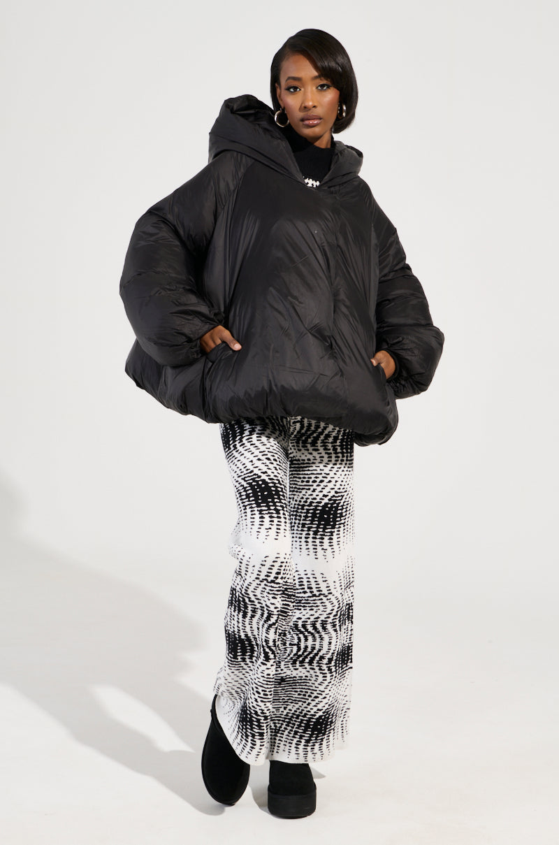 A person dressed in the LEONE Statement Circle Crop Puffer in Black is posing against a plain background, paired with black-and-white patterned pants. They complete their look with black footwear and hoop earrings, keeping their hands tucked into the jacket pockets.