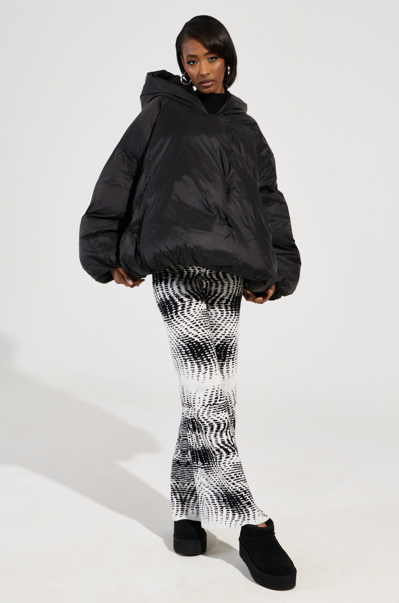 The individual exudes confidence in an oversized LEONE Statement Circle Crop Puffer in Black, complemented by black and white patterned pants and black platform shoes. The plain white backdrop highlights the fashion ensemble.