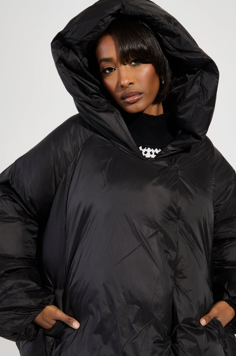 A person is wearing the Leone Statement Circle Crop Puffer in Black, a large and thick hooded coat featuring double-breasted closures. Gazing forward with hands in pockets, the puffy and warm coat is perfect for cold weather. The plain, light-colored background enhances the A-line hem of this stylish outerwear.