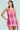 A woman confidently poses in an ELLE TWEED MINI DRESS, a pink plaid sequin mini dress with thin straps, reminiscent of Legally Blonde. With long brown hair, she looks slightly to the side while holding a small black purse against a plain light green background.