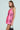 A person with long brown hair wears the ELLE TWEED MINI DRESS in pink, holding a small black handbag by their side. They look over their shoulder against a light blue background, exuding confident vibes reminiscent of 