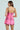 A woman stands facing away, wearing the ELLE TWEED MINI DRESS with black straps and sequin embellishments. Her long dark hair cascades down her back, evoking a Legally Blonde vibe against a soft pastel green background.