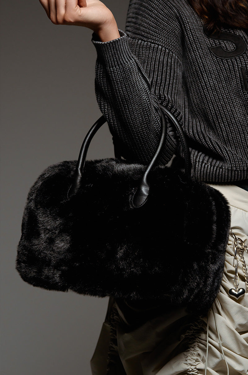A person is showcasing the "TOO FURRY TO CARE" faux fur bag with leather handles. They are wearing a dark ribbed sweater and a light-colored satin skirt adorned with chain details, exuding elegance against the plain, muted backdrop.