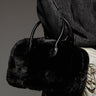 A person is showcasing the "TOO FURRY TO CARE" faux fur bag with leather handles. They are wearing a dark ribbed sweater and a light-colored satin skirt adorned with chain details, exuding elegance against the plain, muted backdrop.